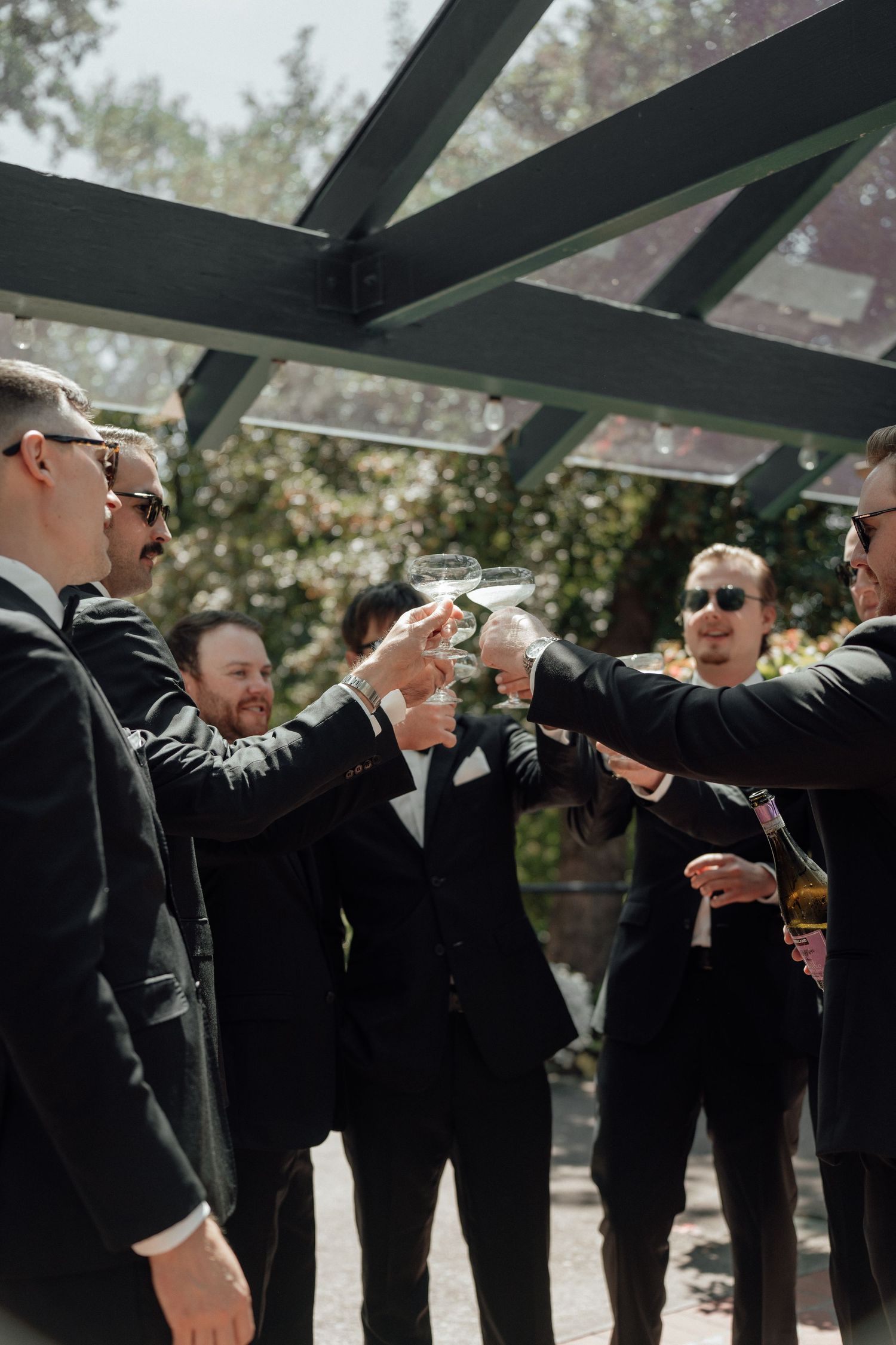 Groomsmen in black suits toast with drinks underneath a wooden pergola at an outdoor wedding celebration.