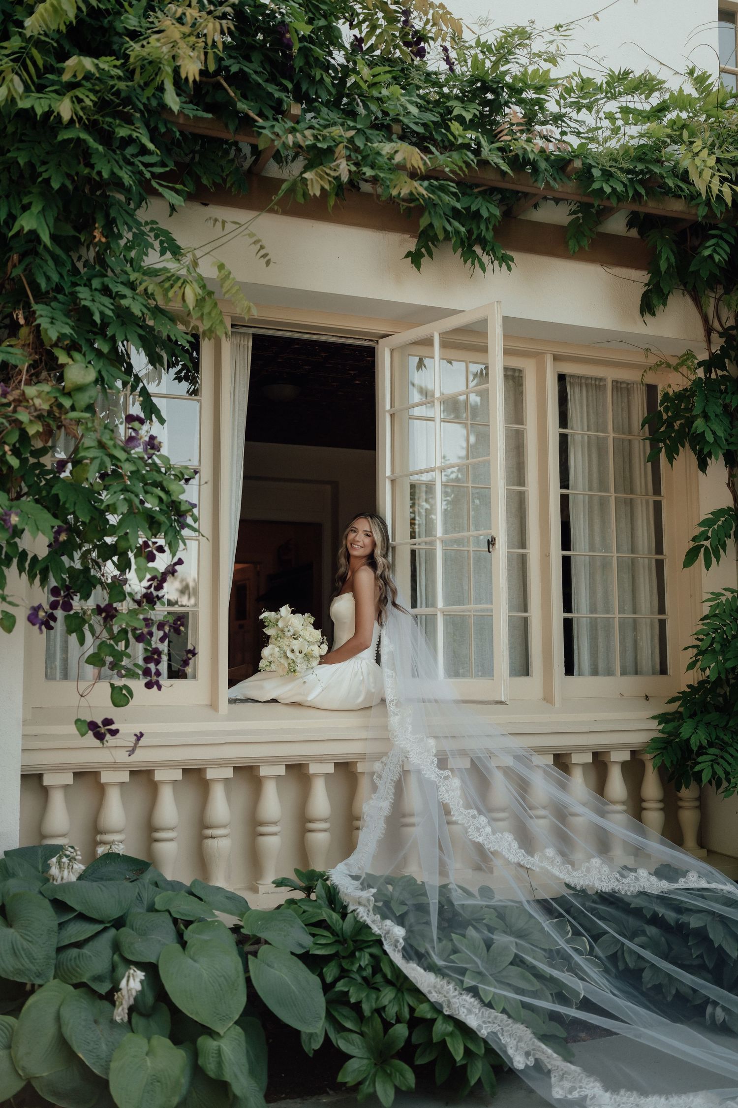A dreamy bridal portrait captured through an ornate window frame with cascading veil and white bouquet.