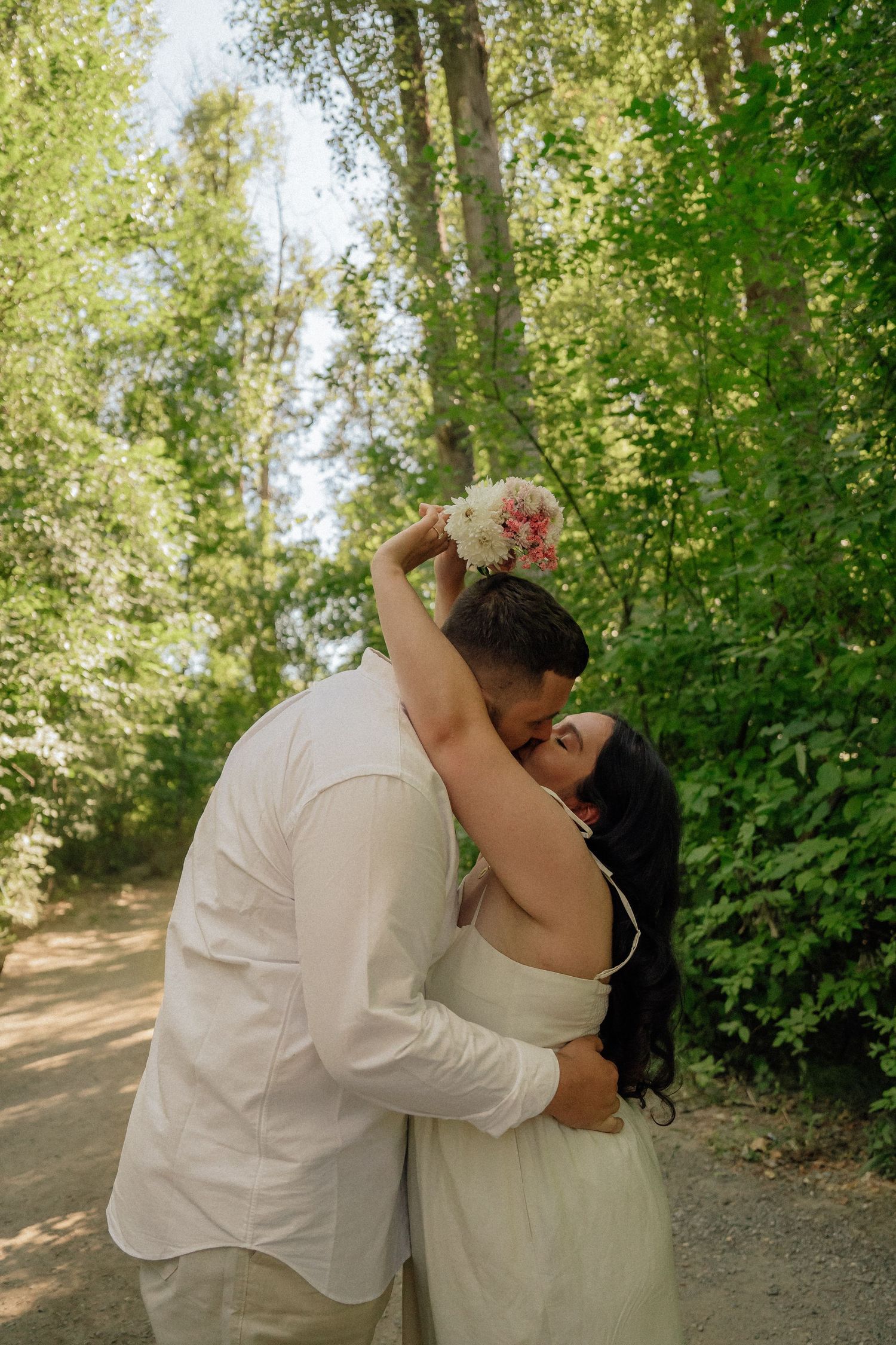 A romantic couple in white embraces on a sunlit forest path surrounded by lush green trees and nature.