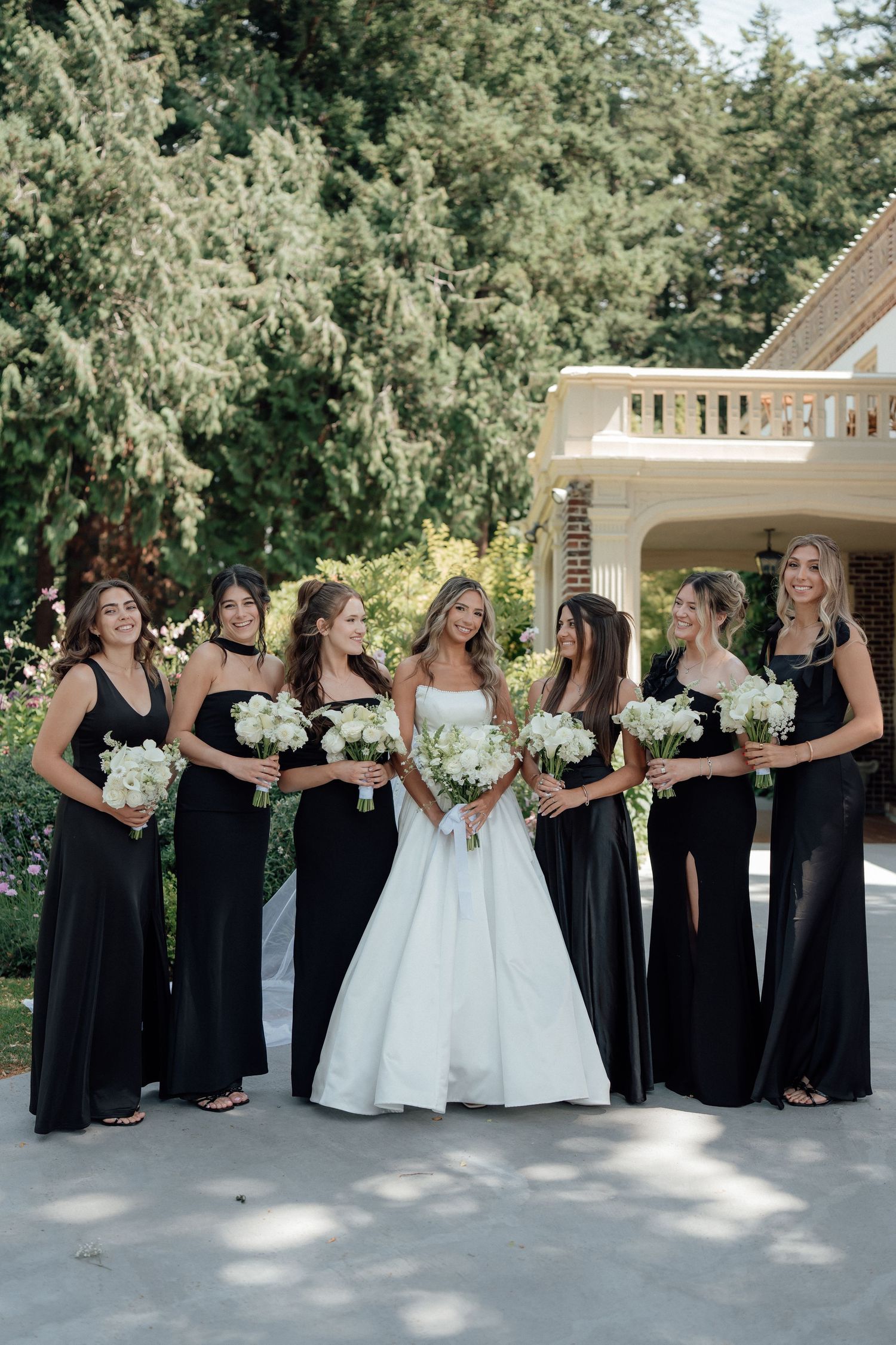 A bridal party poses together wearing elegant black dresses and holding white bouquets.