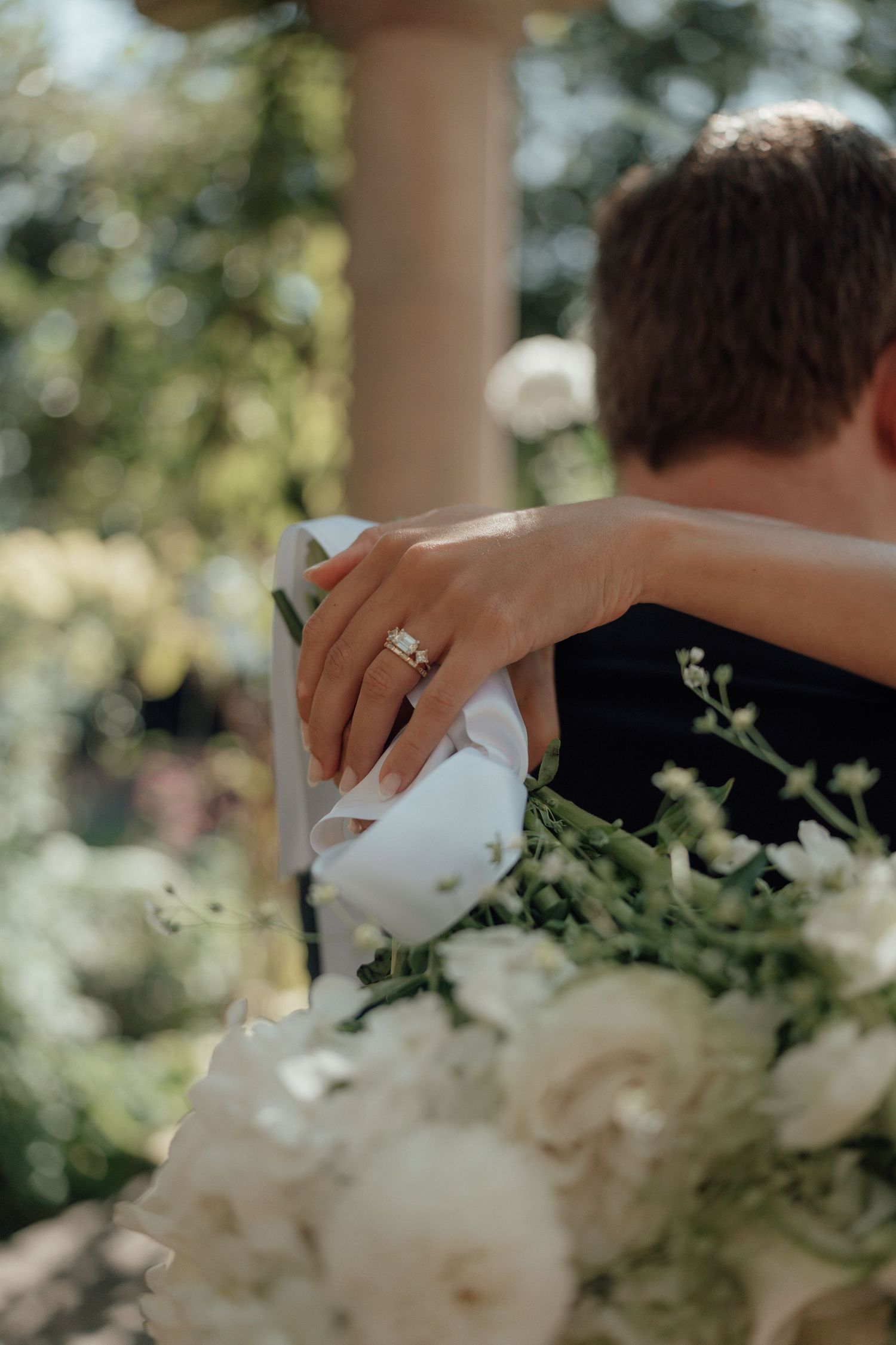 A wedding embrace shows an engagement ring sparkling while white flowers bloom in the soft sunlit background.