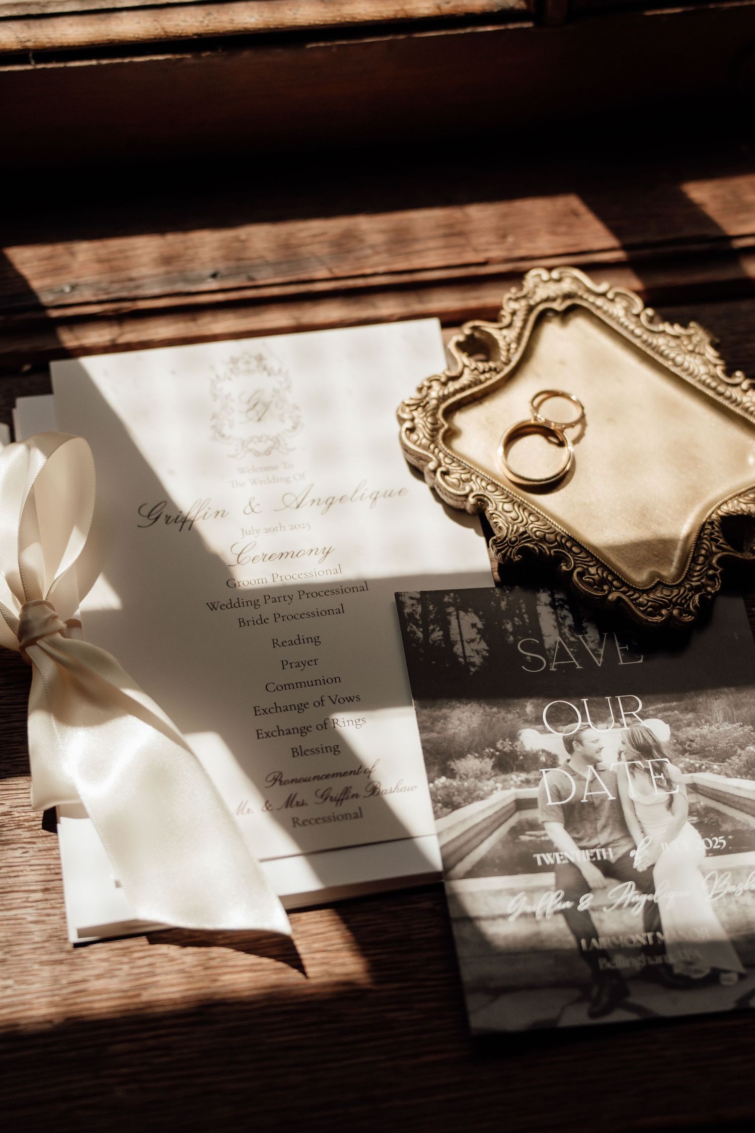 Wedding invitation with white ribbon, save the date card, and gold vintage tray holding rings on wooden surface.