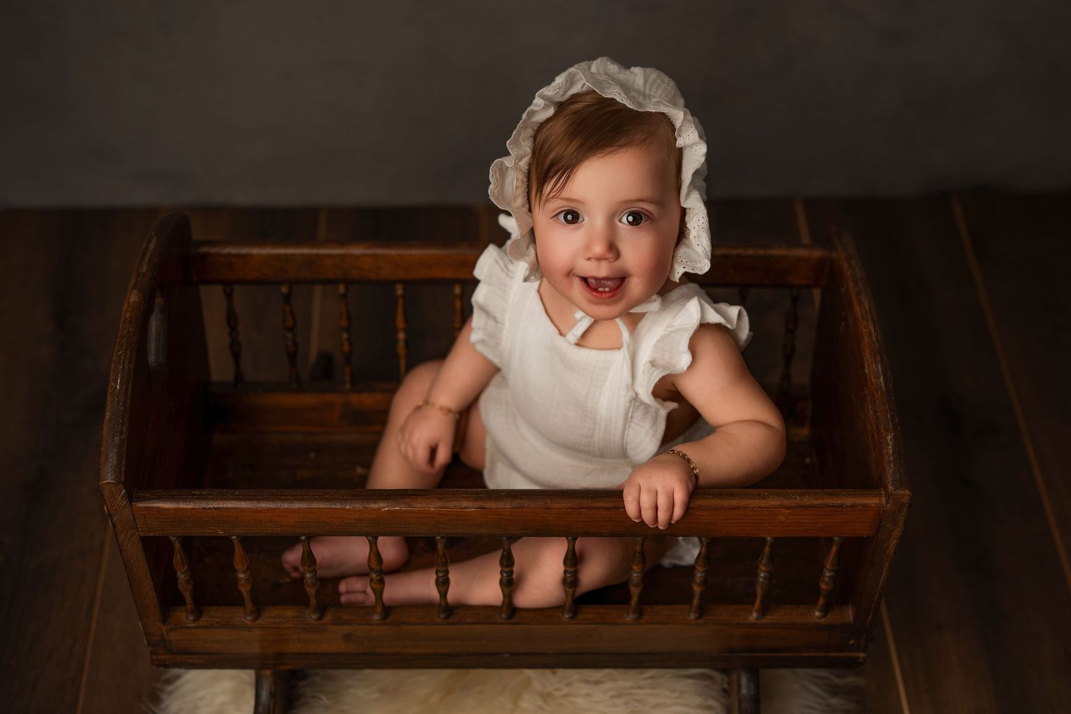A baby in a white dress sits on rustic wooden steps while looking up at the camera.