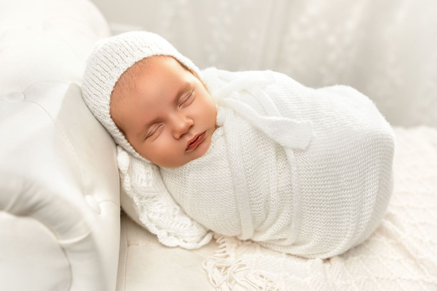 Smiling newborn baby swaddled in white knit wrap and bonnet, sleeping peacefully on white blanket.