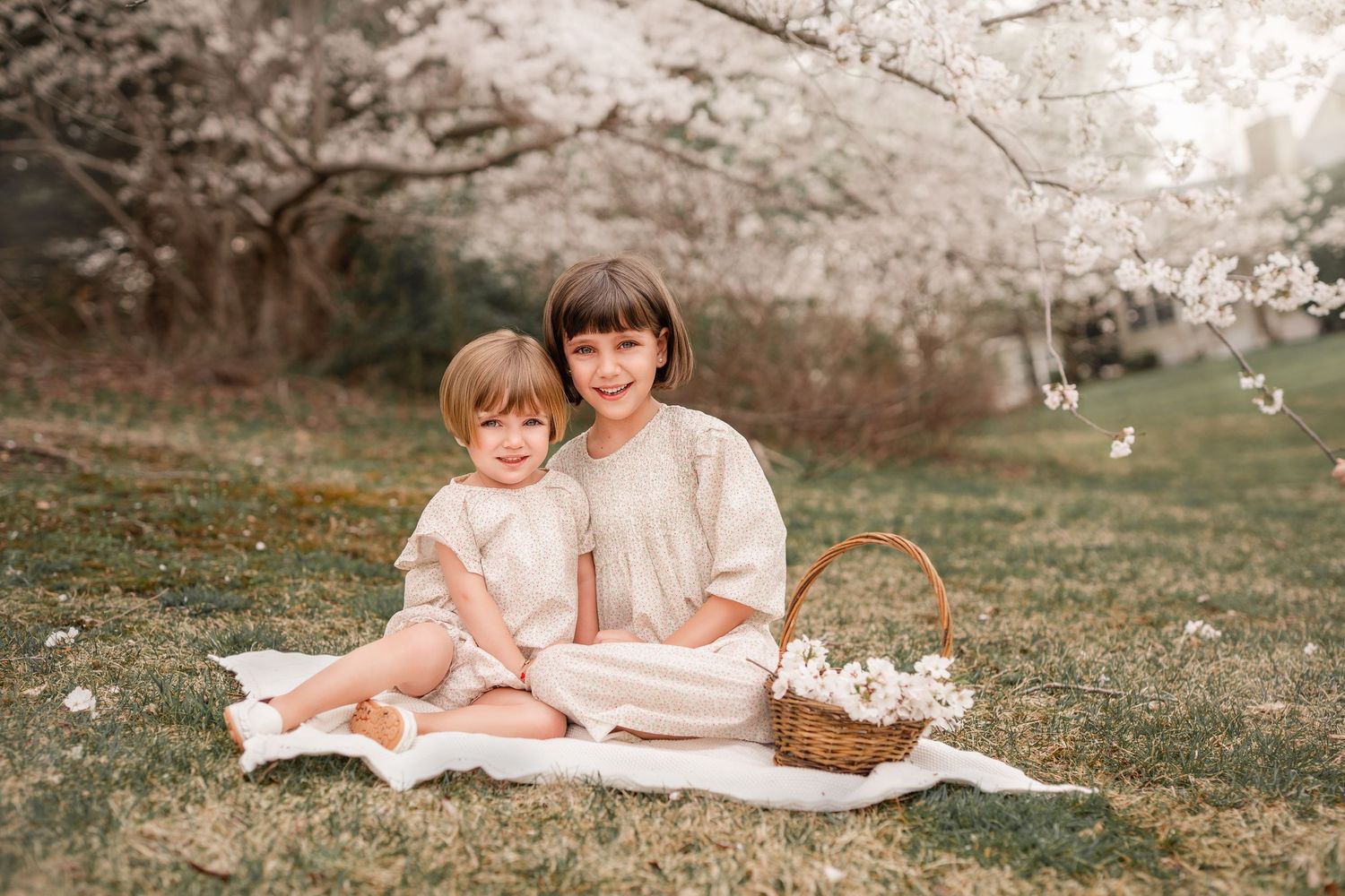 Two young girls in white dresses sit on a blanket under blooming cherry trees with a wicker basket of flowers.