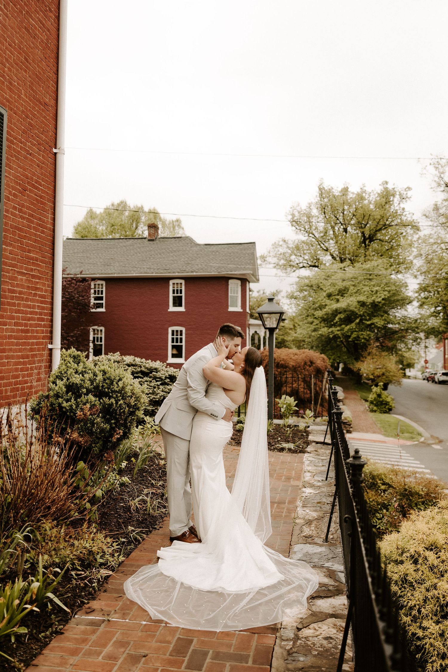 Bride and groom share a romantic dip kiss on a brick path outside a red brick building with lush greenery.
