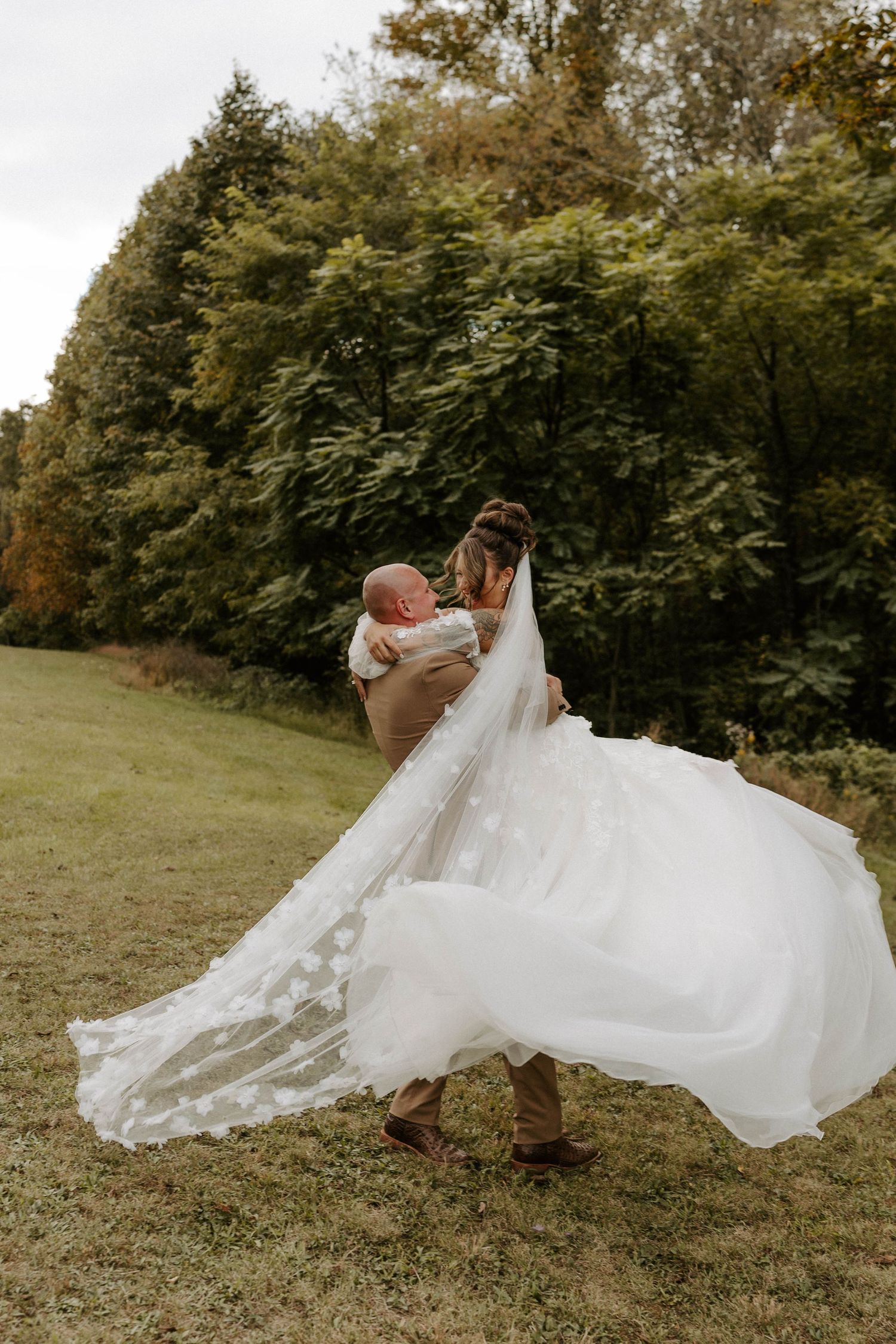 A bride in a flowing white gown and veil is lifted by her groom outdoors on a grassy field with lush green trees.