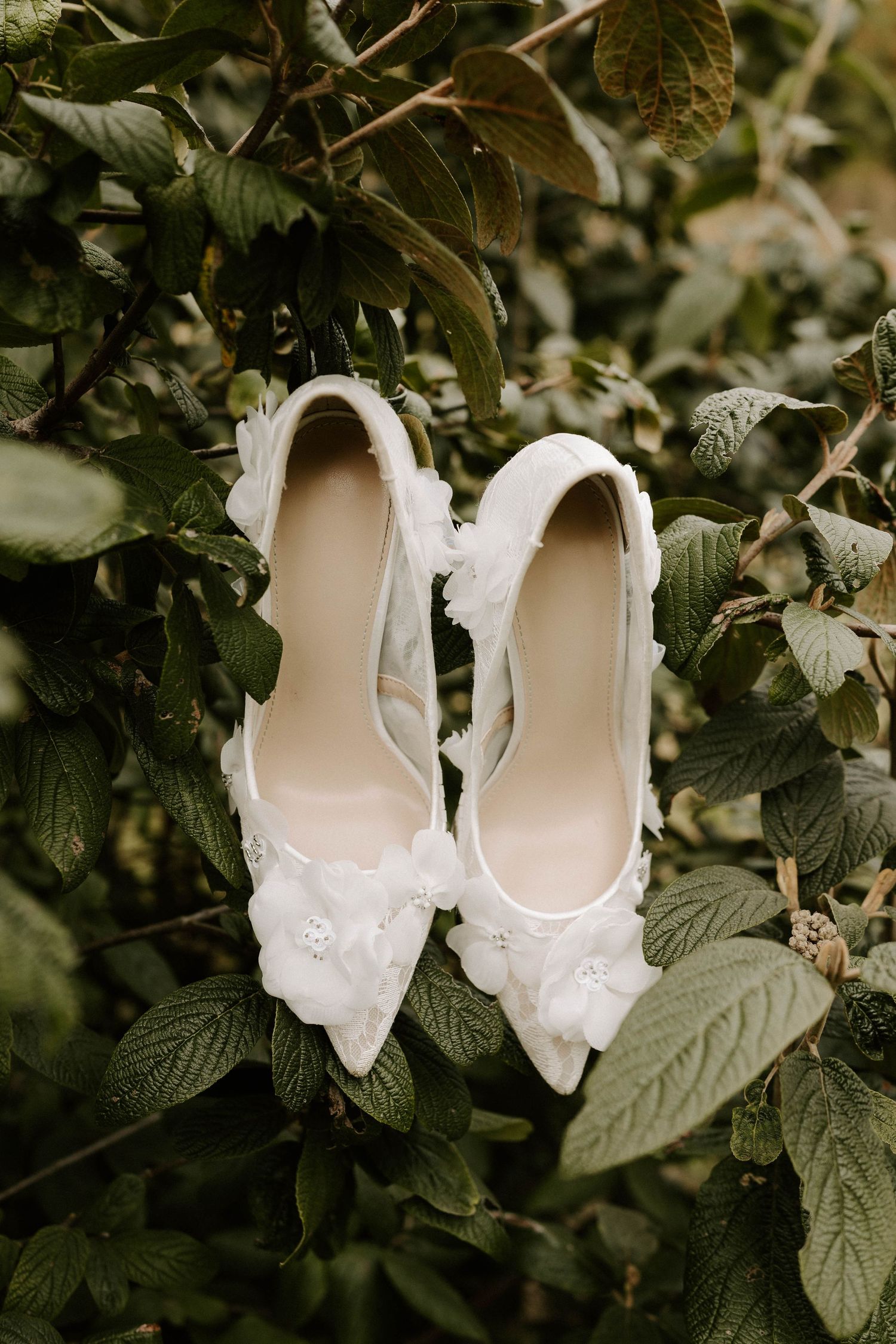 White floral embellished bridal heels displayed among lush green leaves for a romantic wedding detail shot.