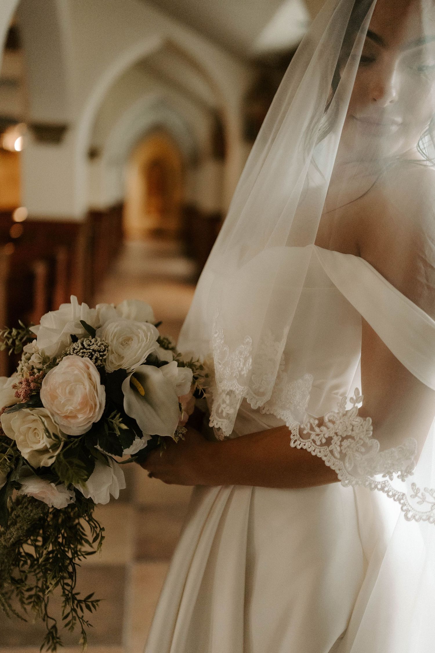 Bride in an off-shoulder satin gown and lace-trimmed veil holds a cream rose and calla lily bouquet in a church aisle.