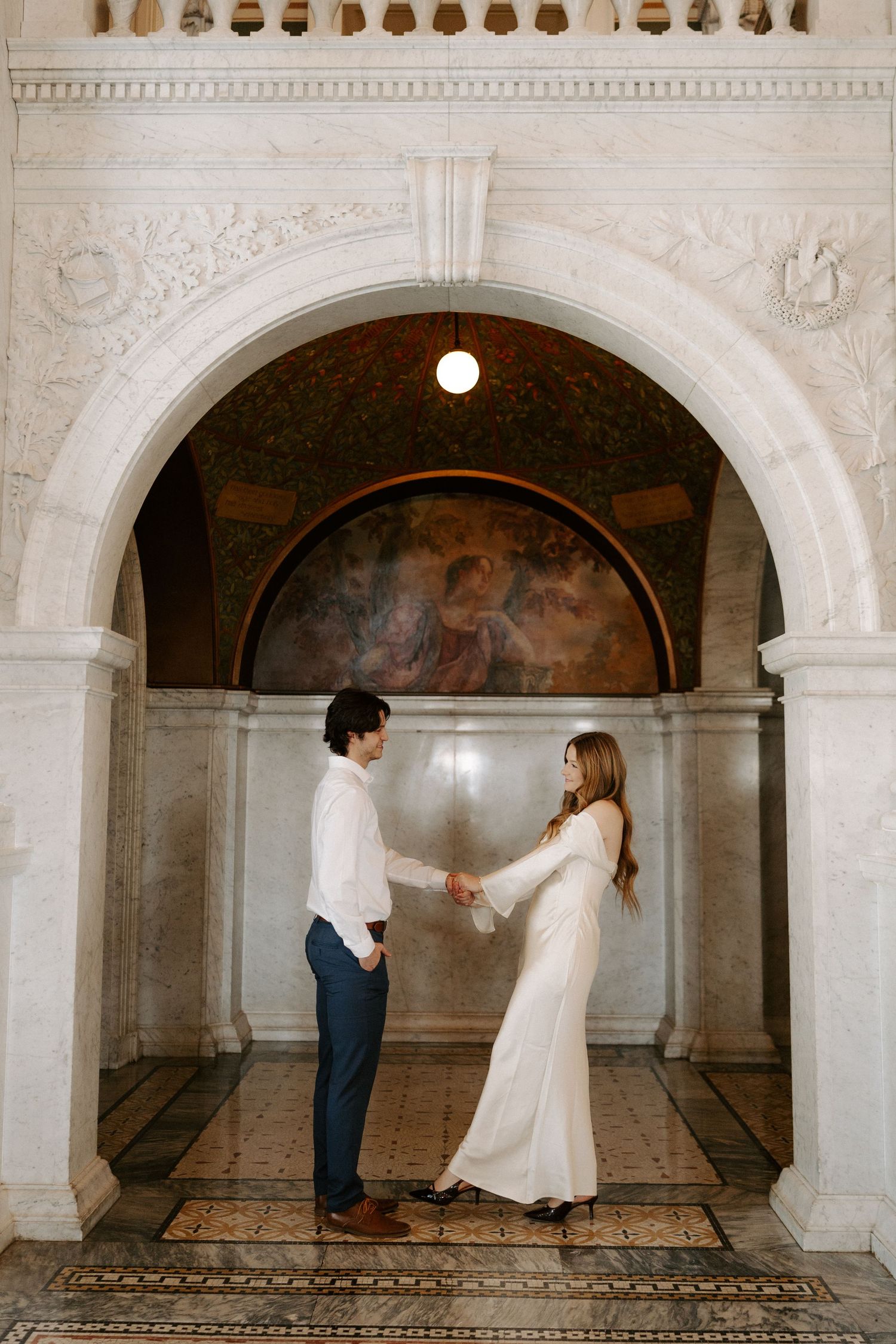 Couple holding hands beneath a grand marble archway with a mural, the bride in a white gown and groom in a white shirt and navy pants.