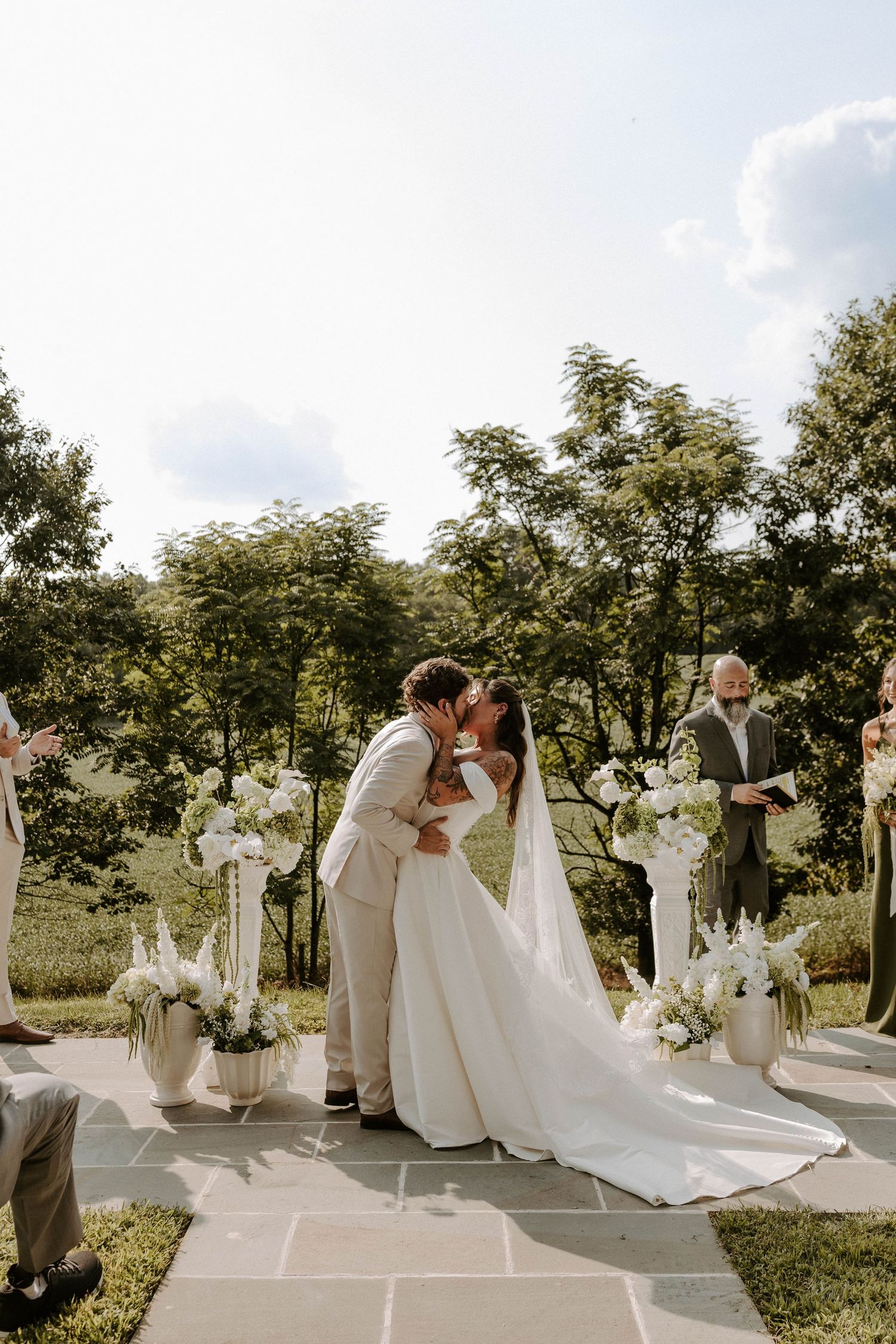 Bride and groom share a kiss during their outdoor wedding ceremony on a stone patio surrounded by white floral arrangements and trees.