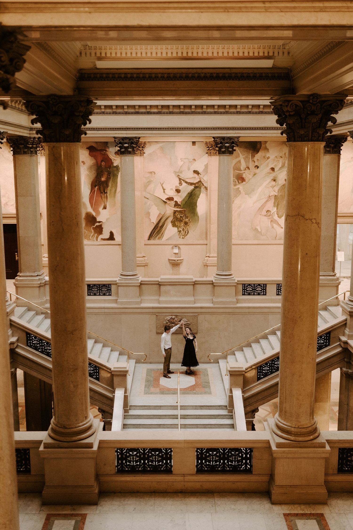 Couple dancing in the center of a grand marble staircase hall with tall columns, ornate railings, and large murals on the walls.