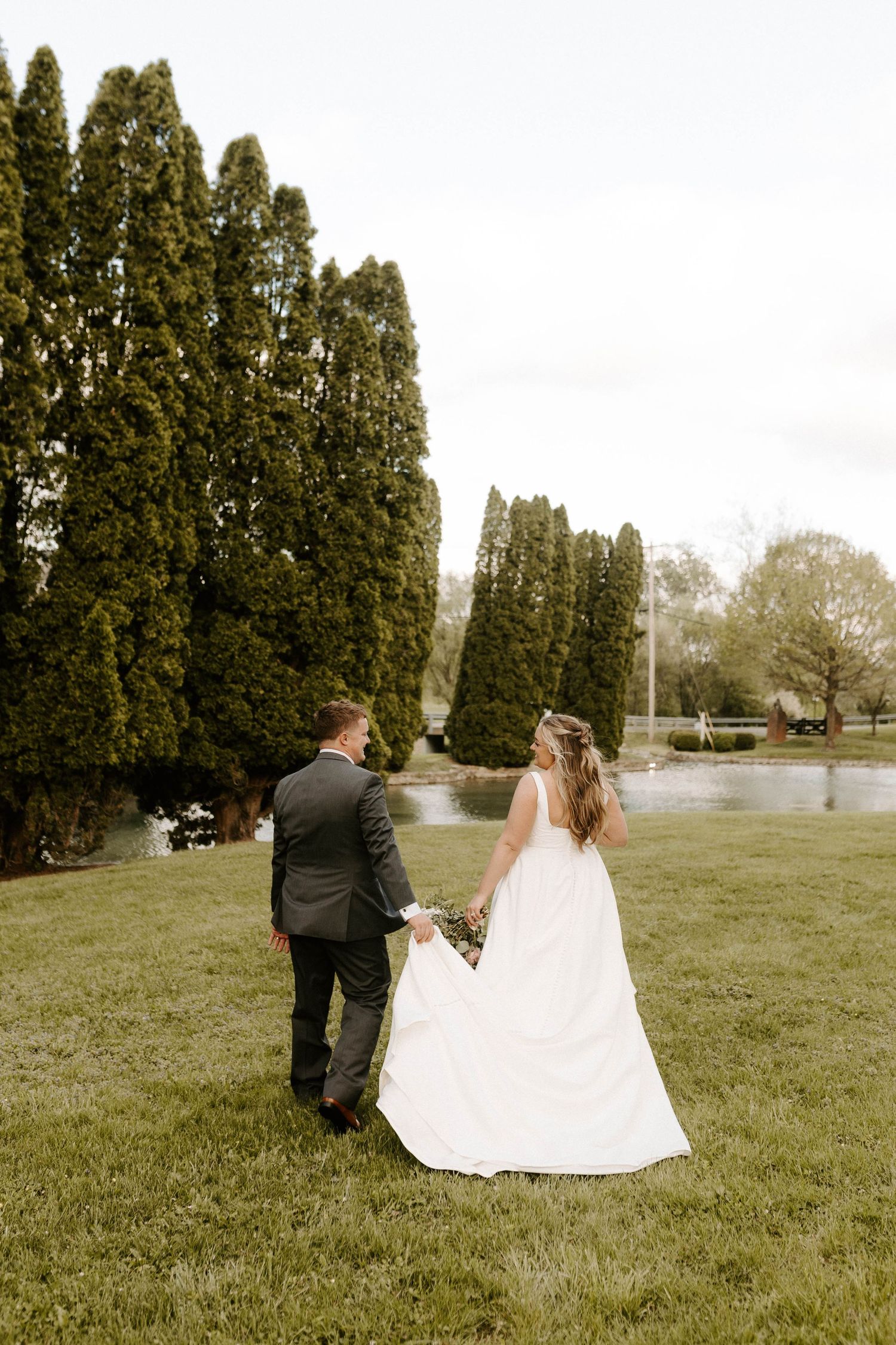 A bride and groom walk hand in hand across a lush green lawn with tall evergreen trees and a pond backdrop.