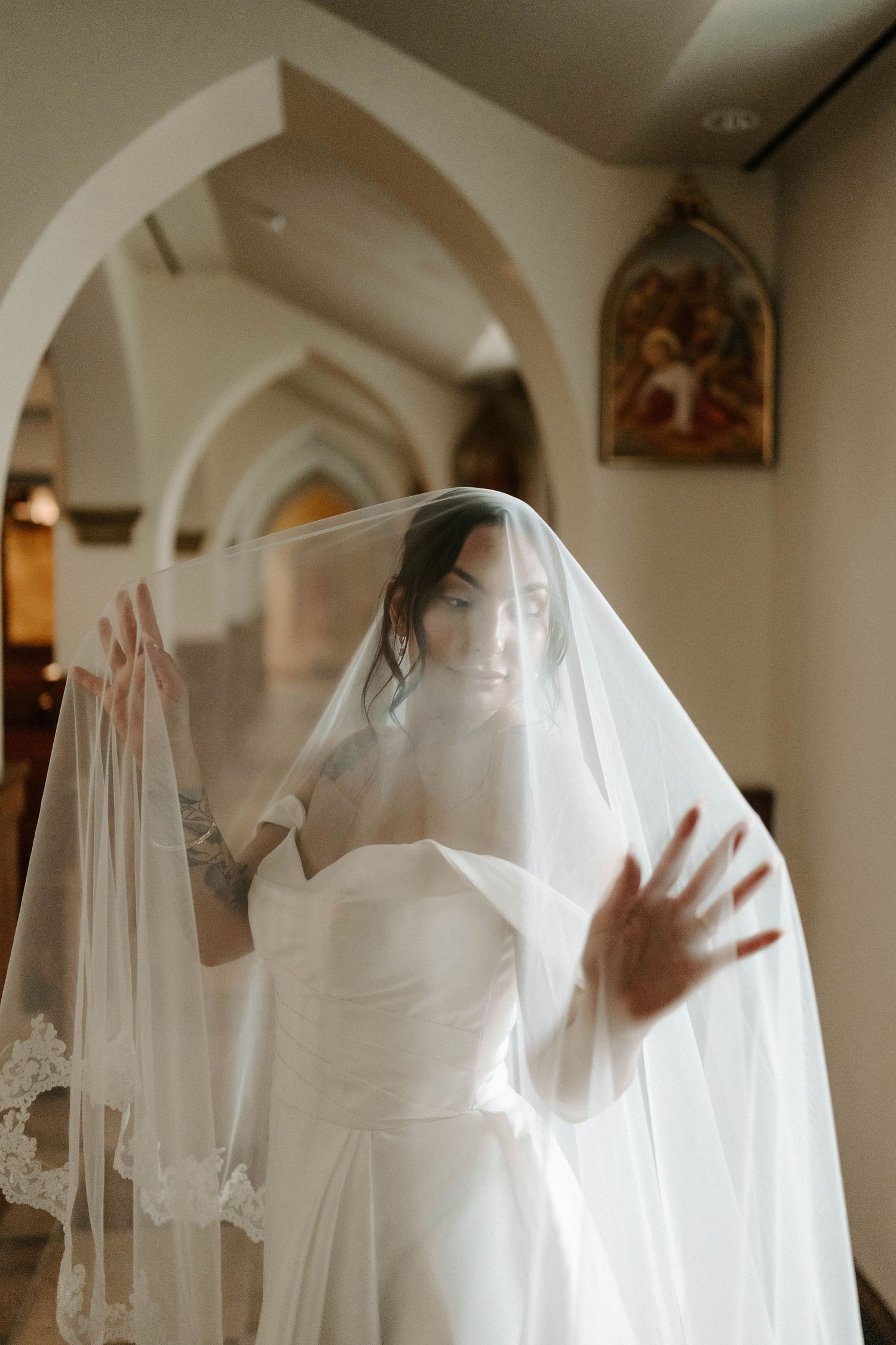 Bride in white strapless gown holds veil over face in elegant church with arched ceilings and religious icons.