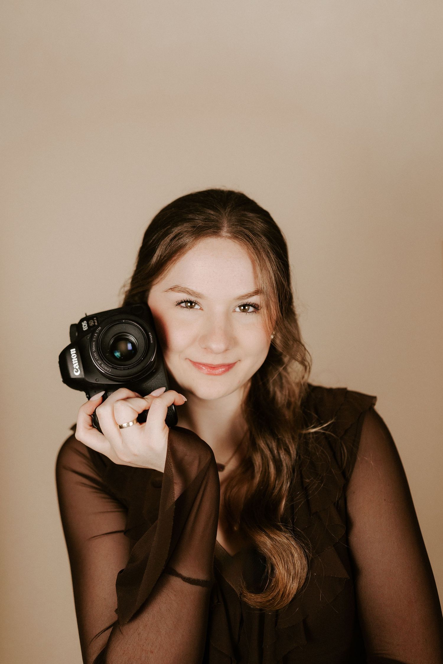 Young female photographer smiling while holding a Canon camera against a neutral beige background.