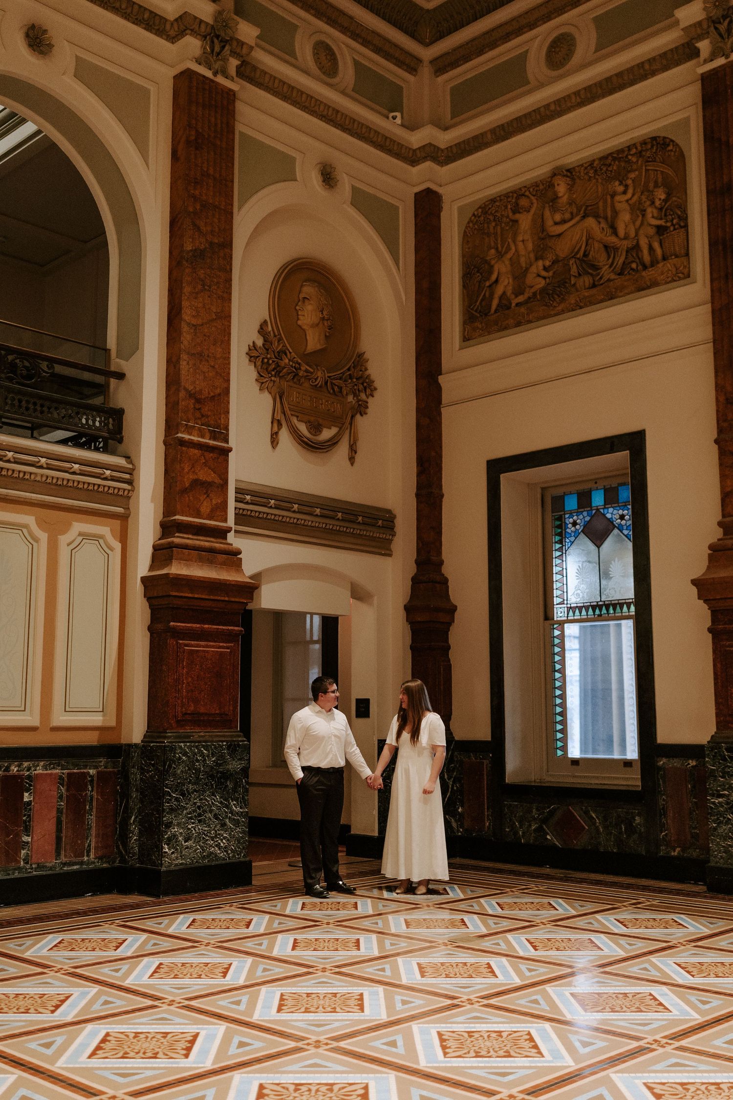 Couple holding hands in an ornate historic hall with marble columns, relief sculptures, stained glass, and a geometric tiled floor.