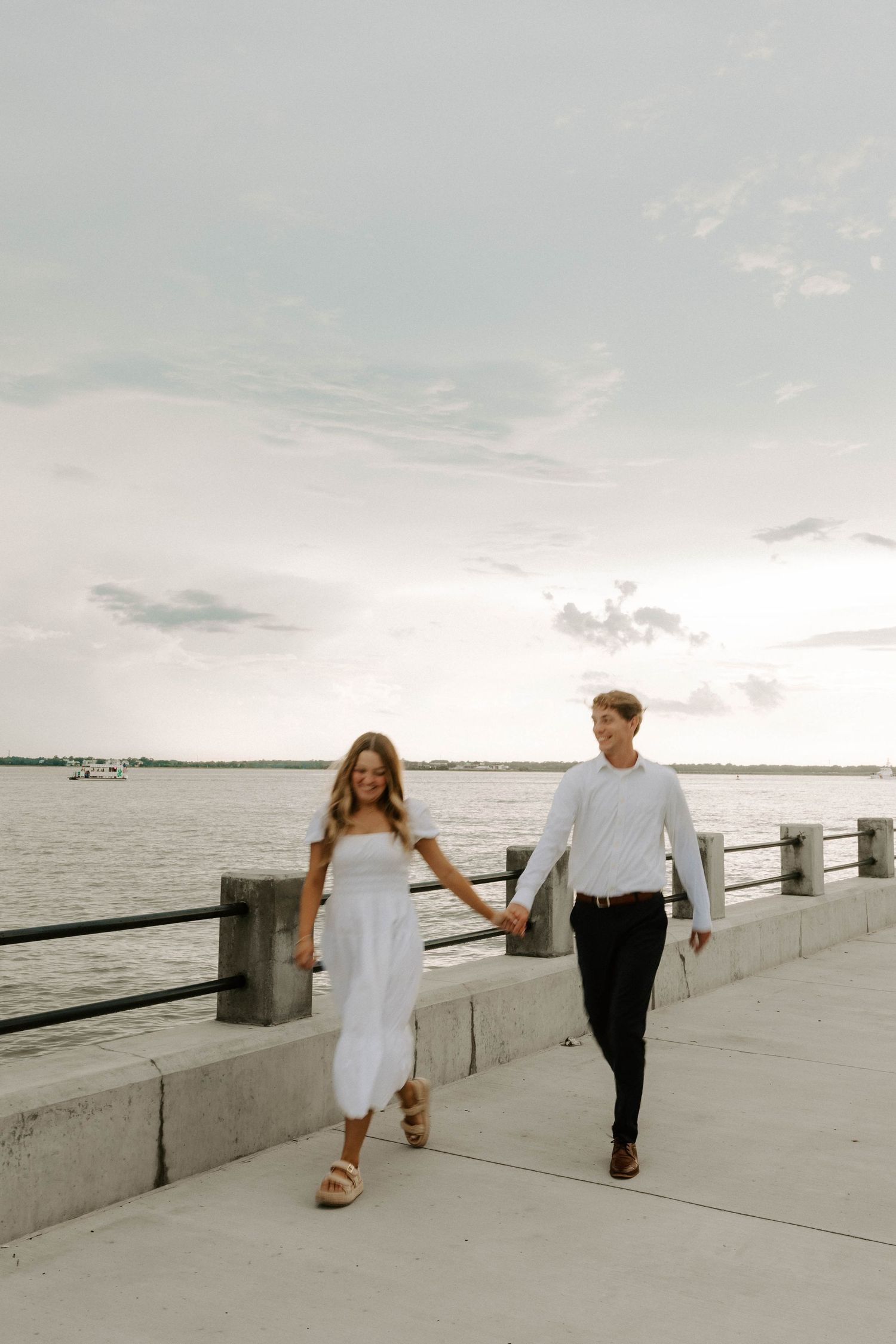 Couple holding hands and walking along a waterfront promenade beside a railing, with calm water and a soft cloudy sky behind them.