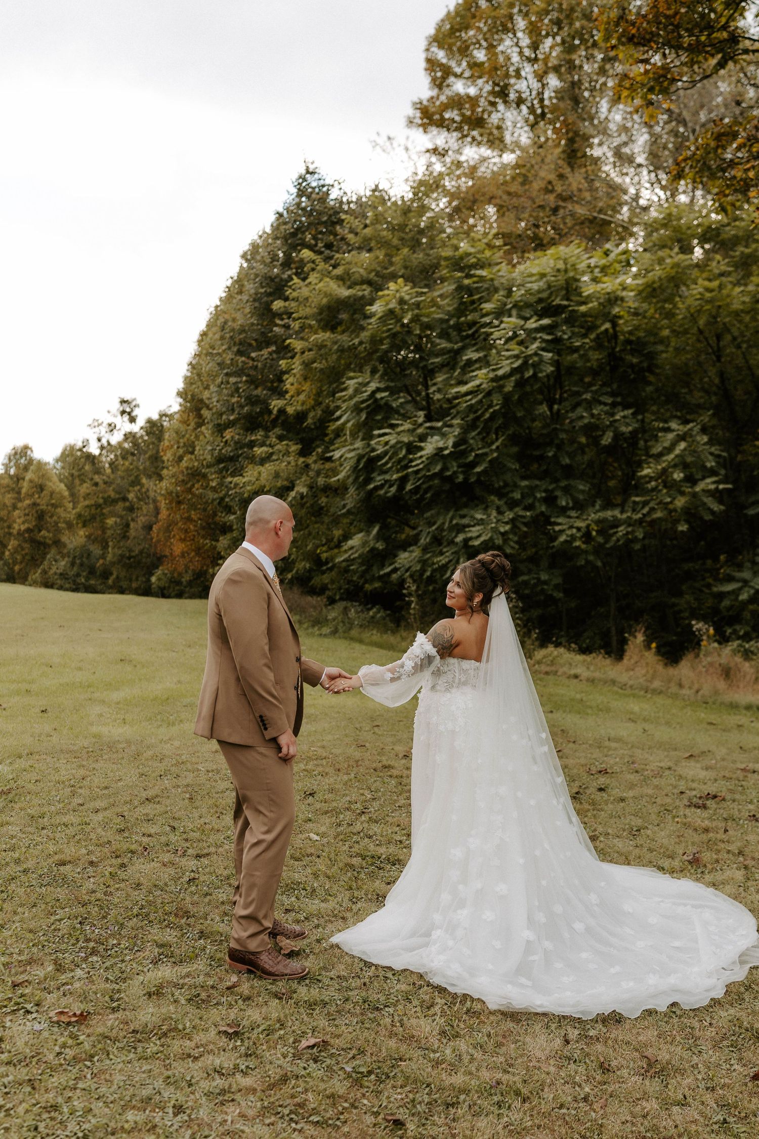 Bride in white lace gown with veil holds groom's hand in tan suit on green meadow with autumn trees backdrop.