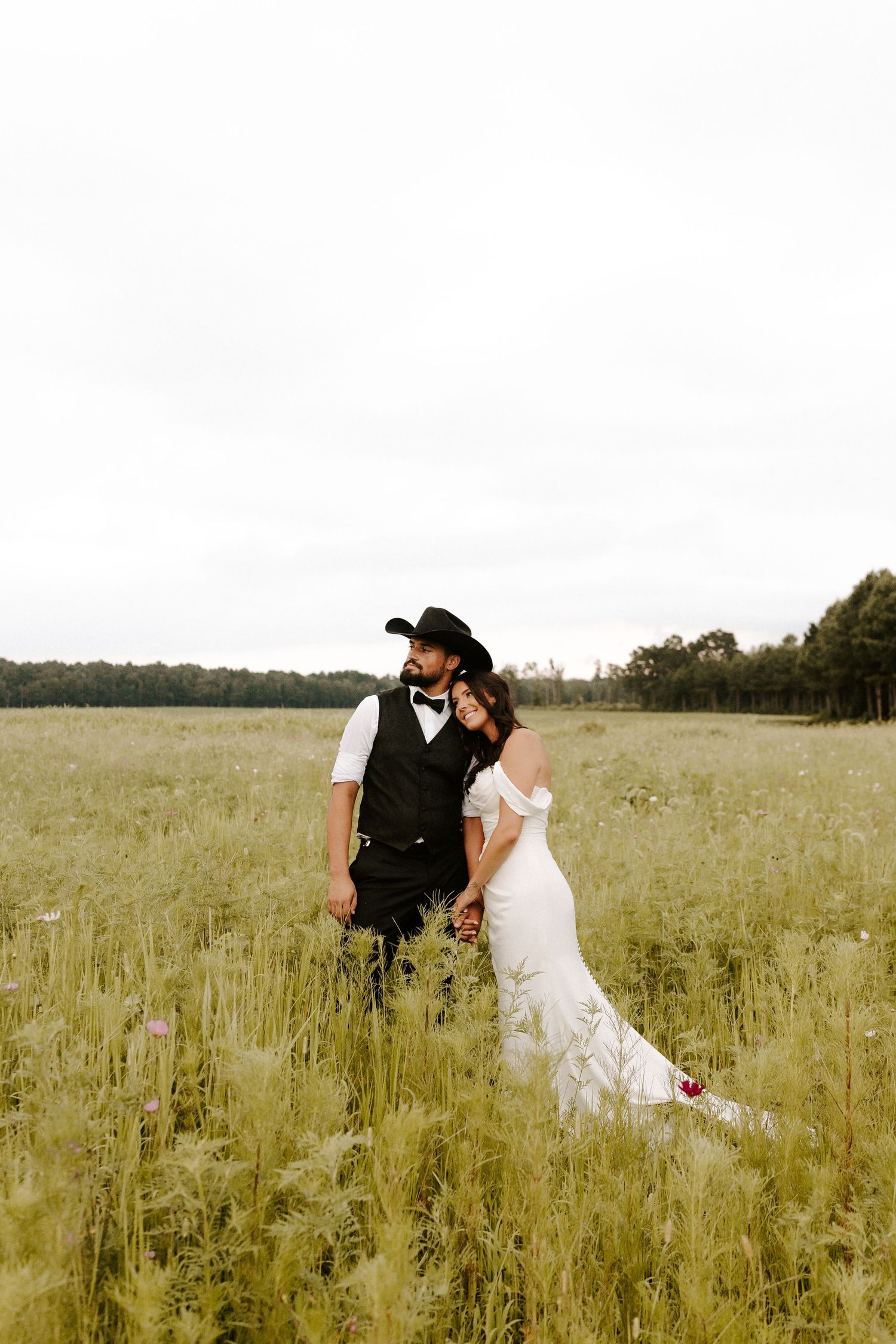 Bride and groom stand together in a lush green field, groom wearing a black cowboy hat and vest.