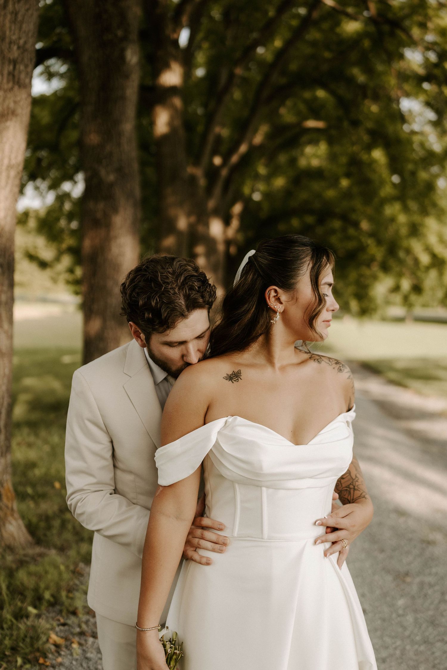 Groom in beige suit kissing bride's shoulder in white off-shoulder gown outdoors among lush green trees.