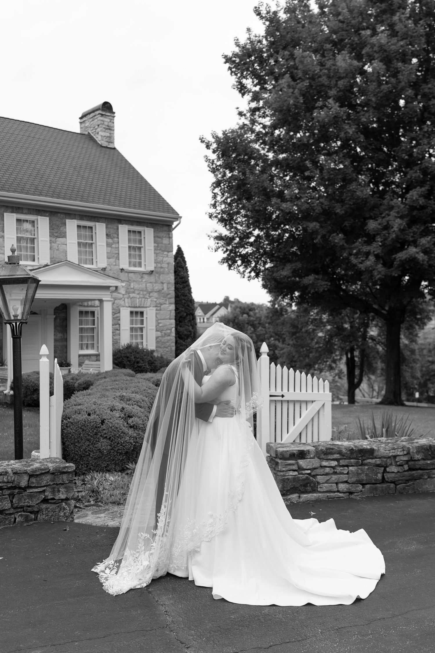 Black and white photo of bride and groom kissing near a stone estate with a white picket fence.