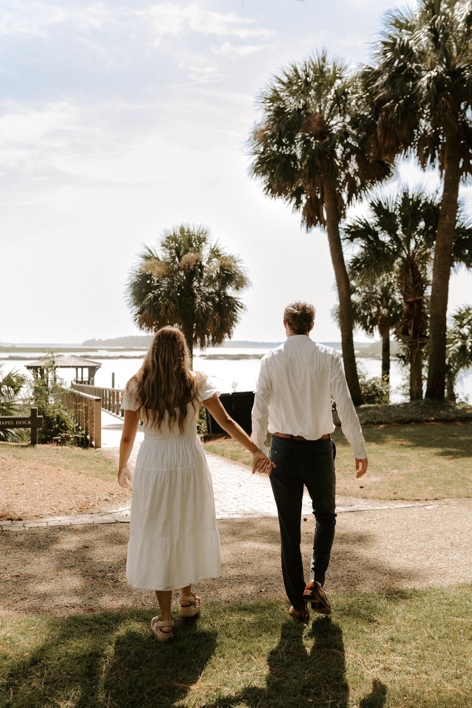 Couple holding hands walking toward a waterfront path lined with palm trees on a sunny day, the woman in a white dress.