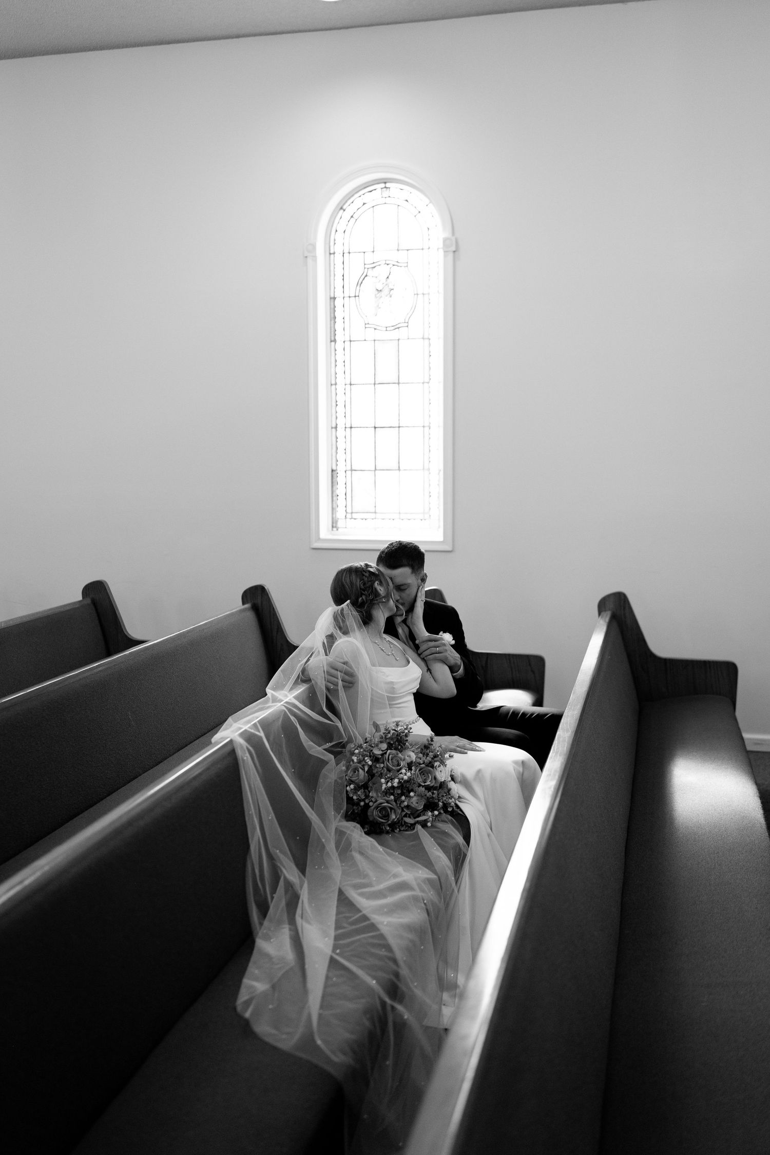 Black and white photo of a bride and groom sharing a kiss in a church pew, with a stained glass window behind them.