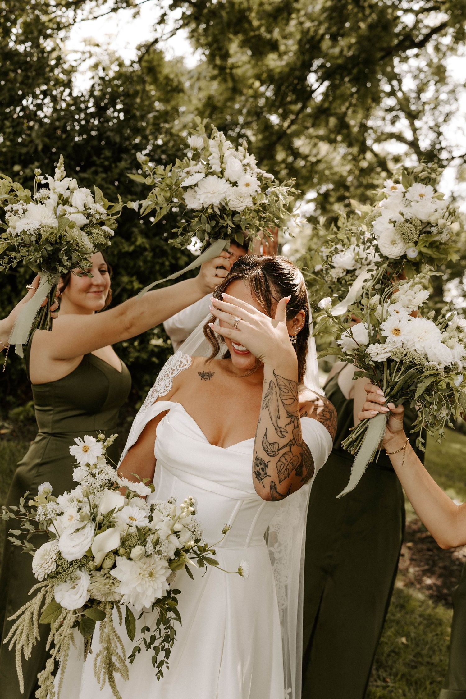 Bride in white gown holds floral bouquet surrounded by bridesmaids in olive dresses with white flower bouquets outdoors.