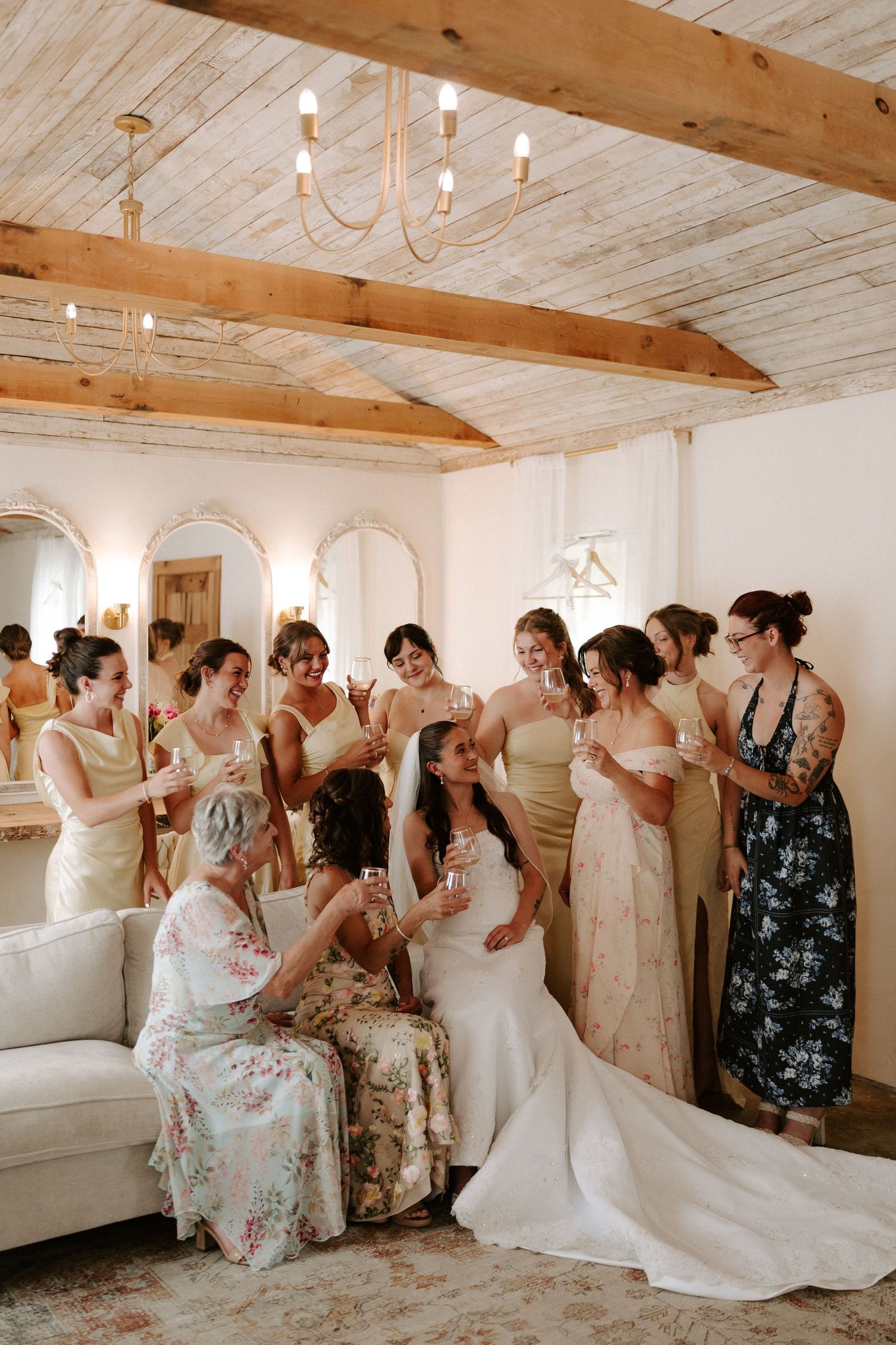 Bride in white gown surrounded by bridesmaids in champagne dresses toasting in a rustic room with wood beam ceiling.