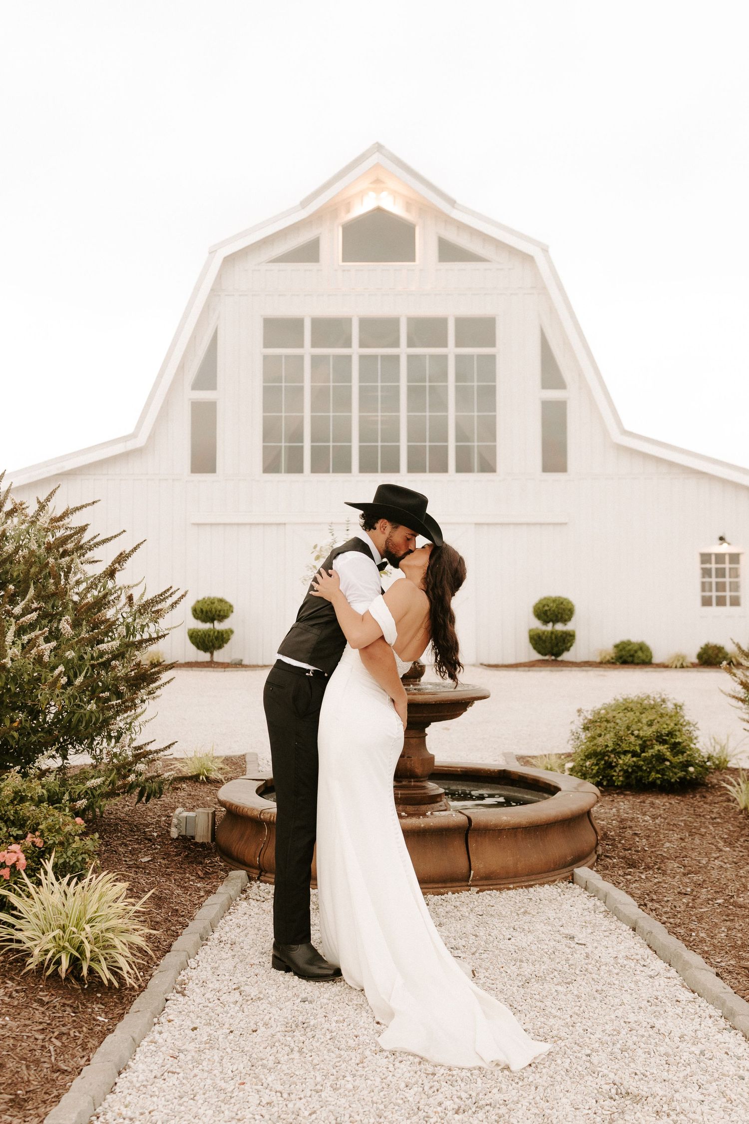 Bride and groom share a romantic kiss in front of a white barn venue, surrounded by lush greenery and a stone fountain.