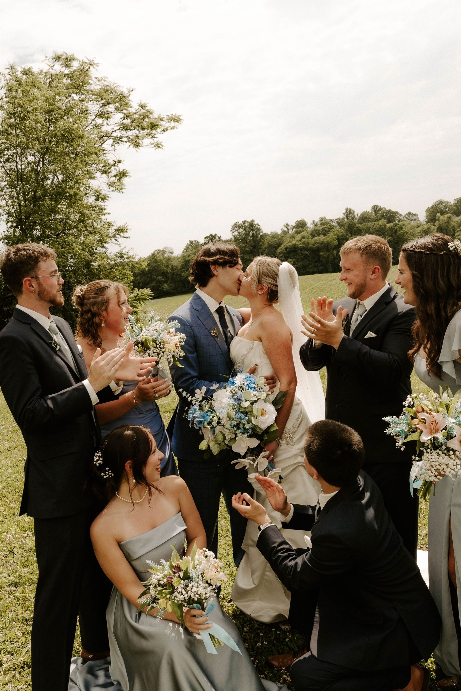 A bride and groom share a kiss surrounded by their wedding party outdoors, with bridesmaids in sage green holding floral bouquets.