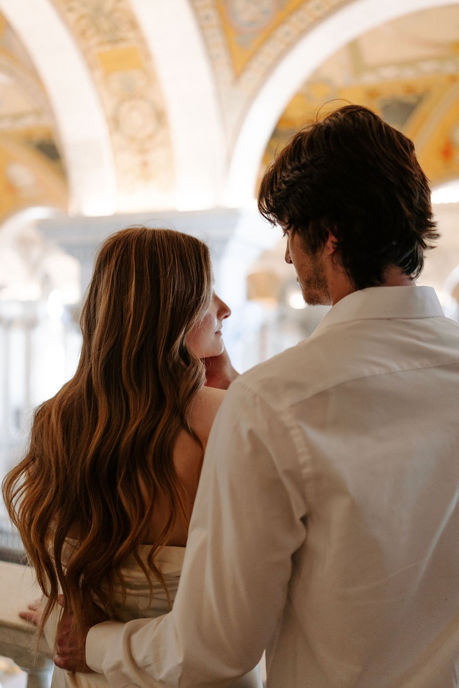 Couple embracing from behind beneath ornate gilded arched ceilings, the woman with long wavy hair and the man in a white shirt.