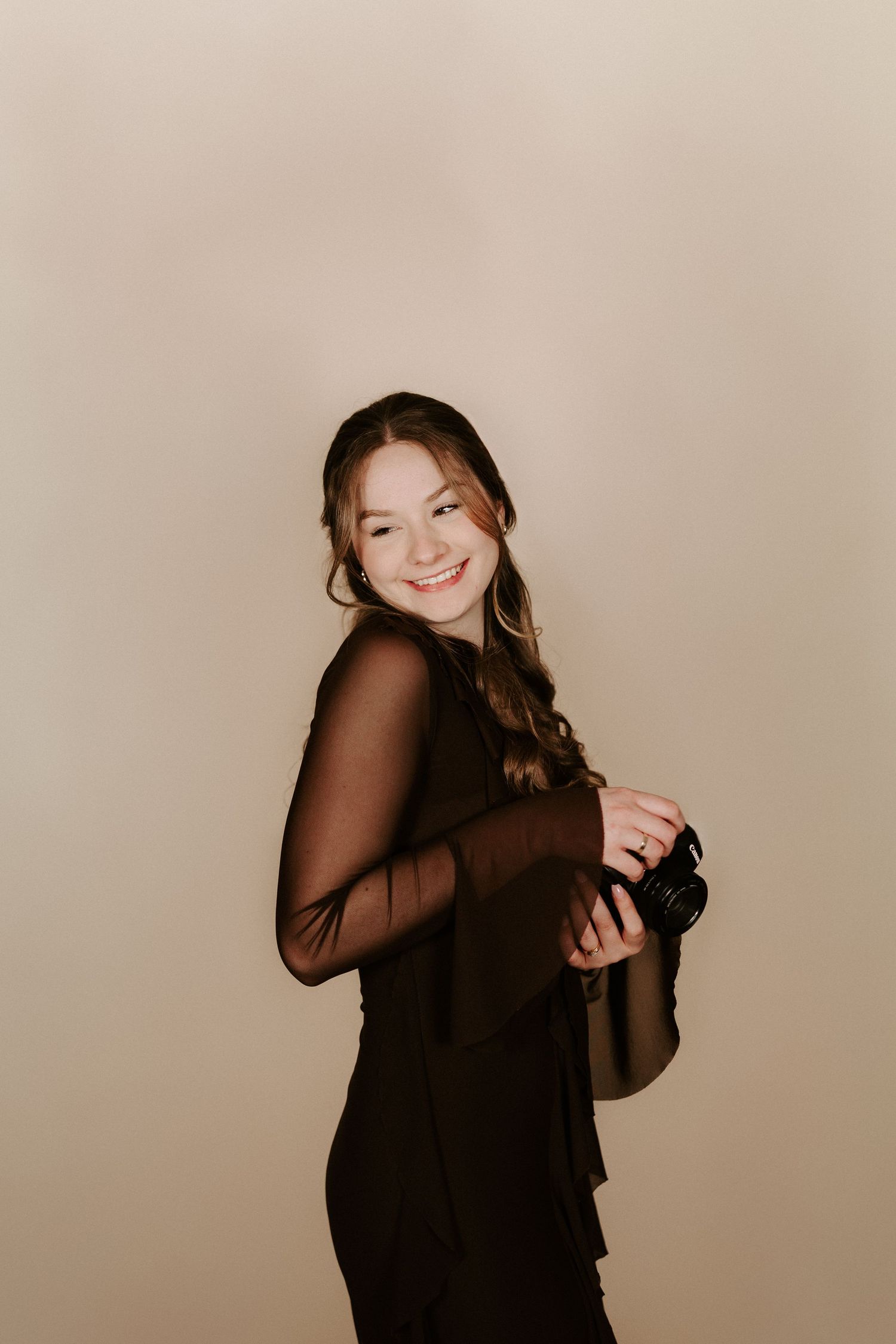 Smiling young female photographer in black outfit holding camera against a warm beige background.