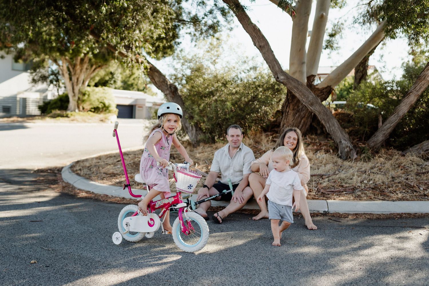 A mum and dad sitting on the kerb of their street, watching their daughter ride a bike with training wheels, and their toddler son running