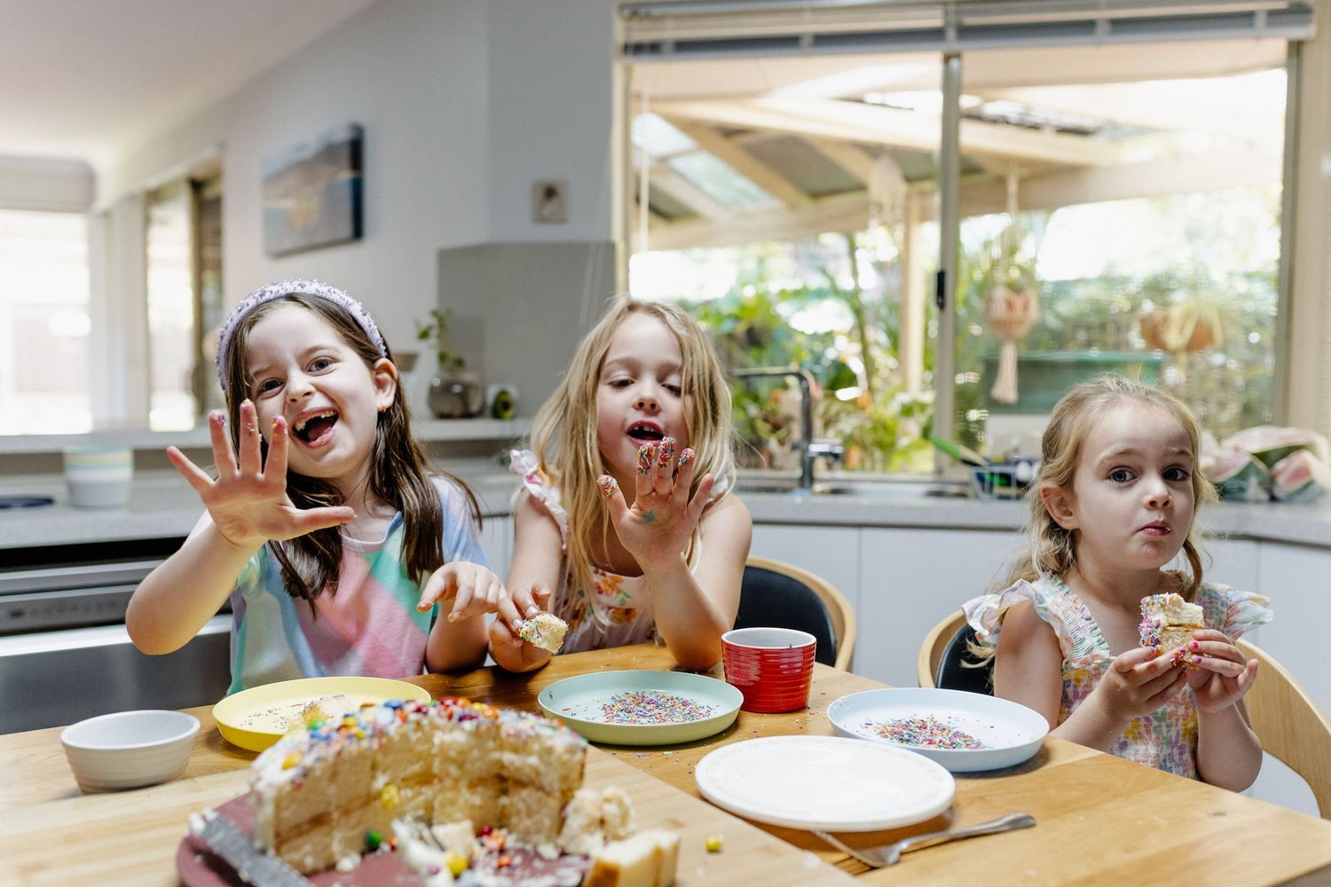 Three young girls sit at a kitchen bench, with cake and cake decorations on their fingers, holding their hands up.