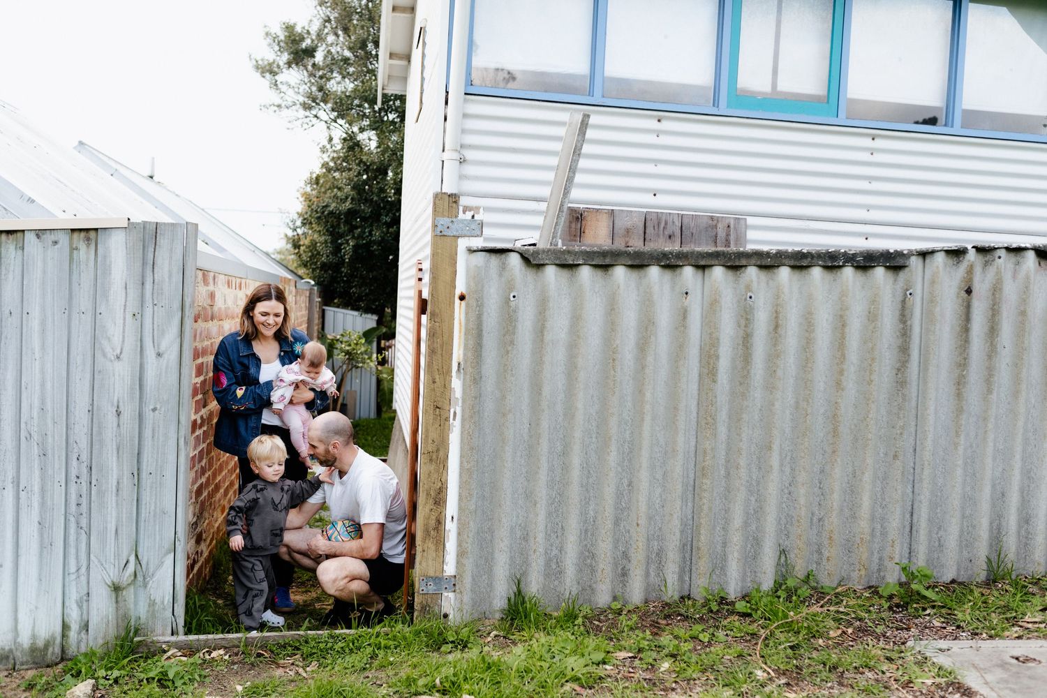 A mum, dad, toddler son, and baby daughter, standing and sitting in their gateway, framed by the back fence of their house