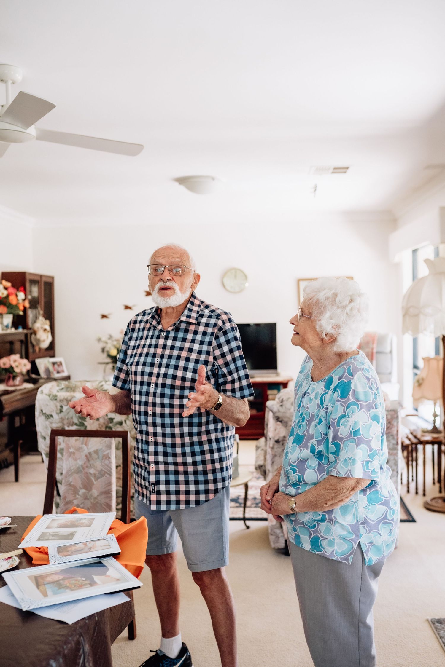 An elderly man and woman stand next to a table with old photos, telling stories.