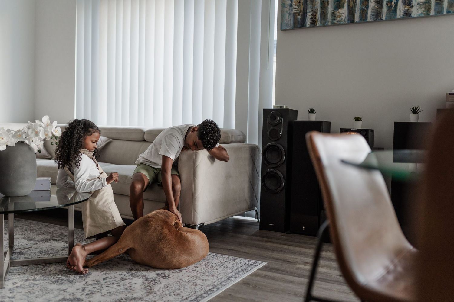 A South African boy and girl sit on the floor of their lounge room. The boy pats their tan dog, and the girl watches.