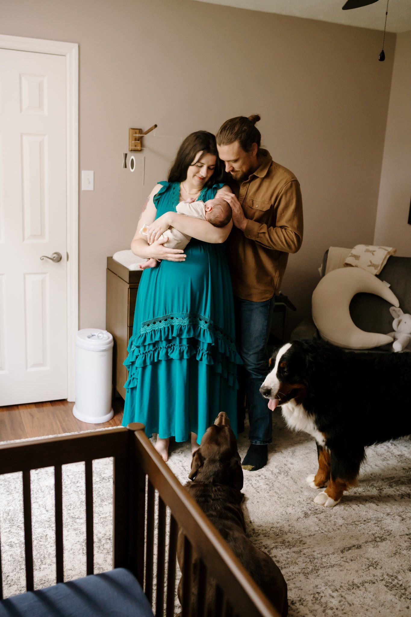 Parents stand in nursery holding newborn while family dogs run around the nursery