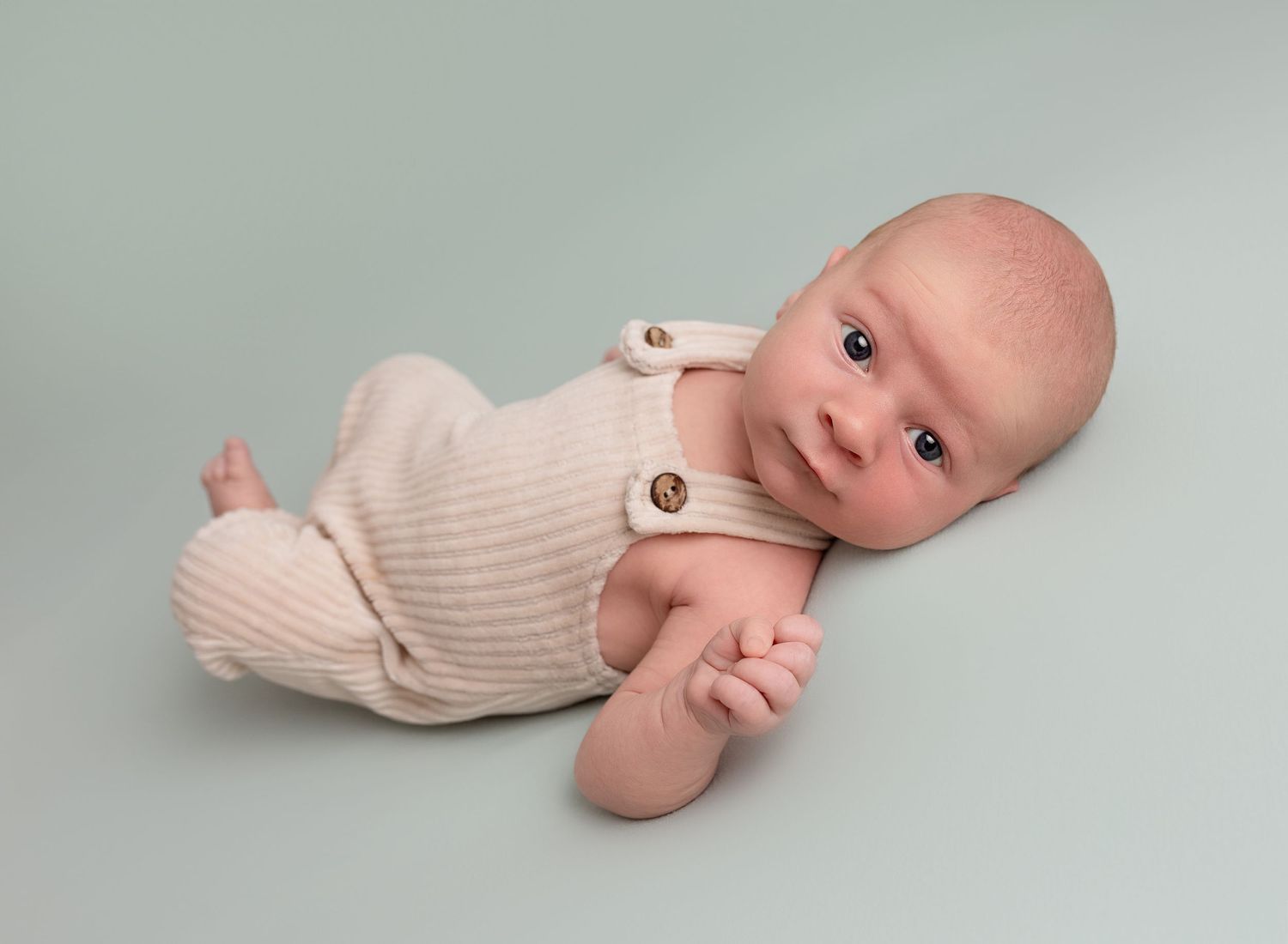 Baby boy looking at camera wearing corduroy dungarees on a pale sage green backdrop