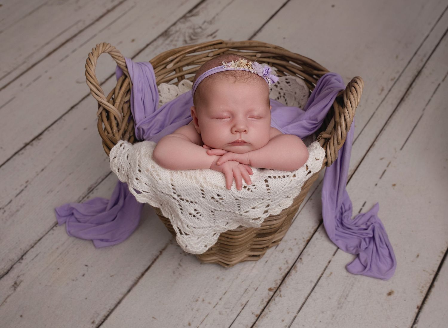 Baby girl with chin on hands leaning forward in wicker basket, lilac accents. newborn photoshoot