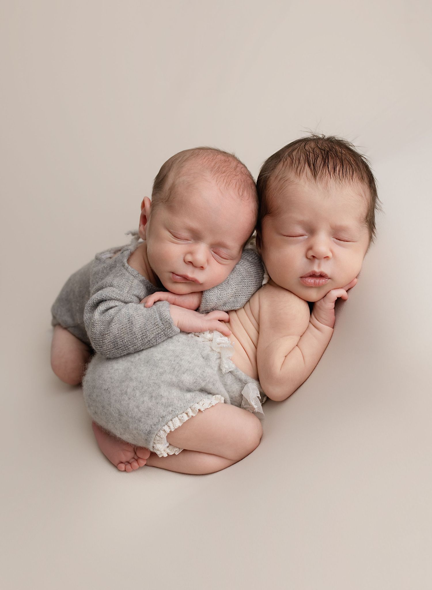 Newborn boy-girl twins, both wearing grey outfits at their professional photoshoot. Boy is posed gently on top of girl and both are sleeping