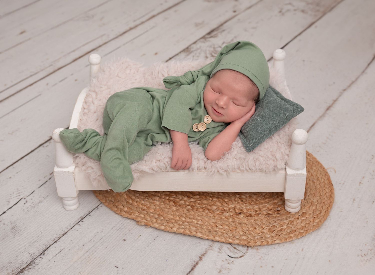 Newborn boy asleep on small wooden bed wearing green sleep suit and hat, jute rug on white wooden floor. Baby photography, Renfrewshire