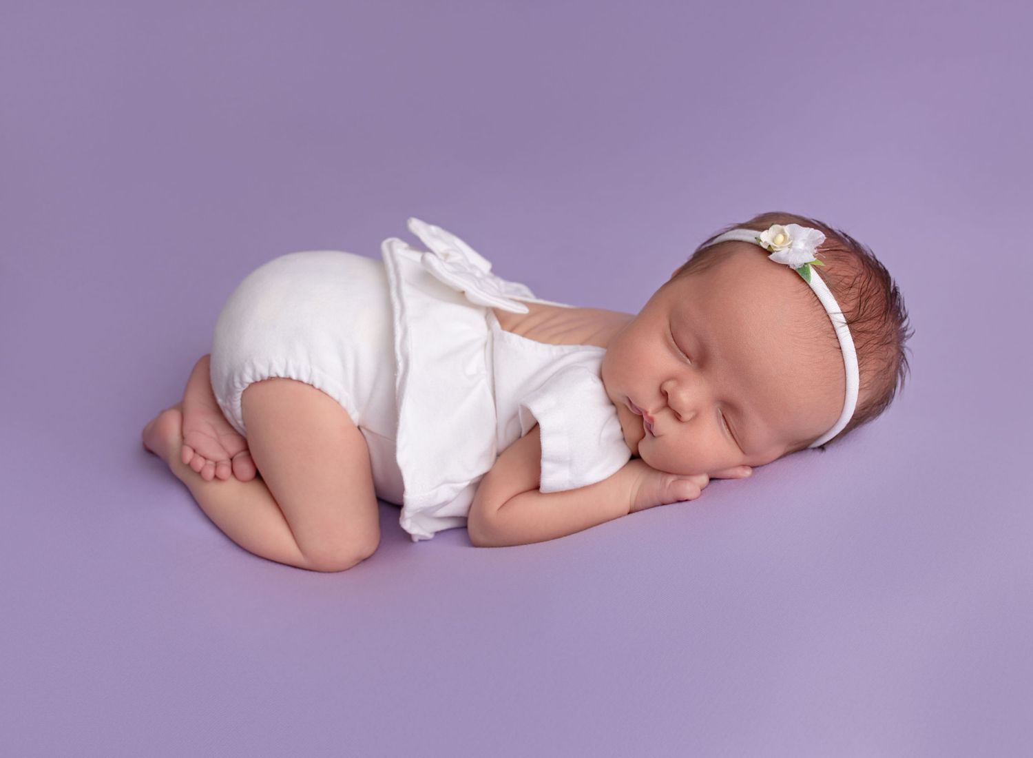 New baby girl asleep on tummy, bum up pose wearing white dress and headband, lilac background. Newborn photoshoot Glasgow, Renfrew
