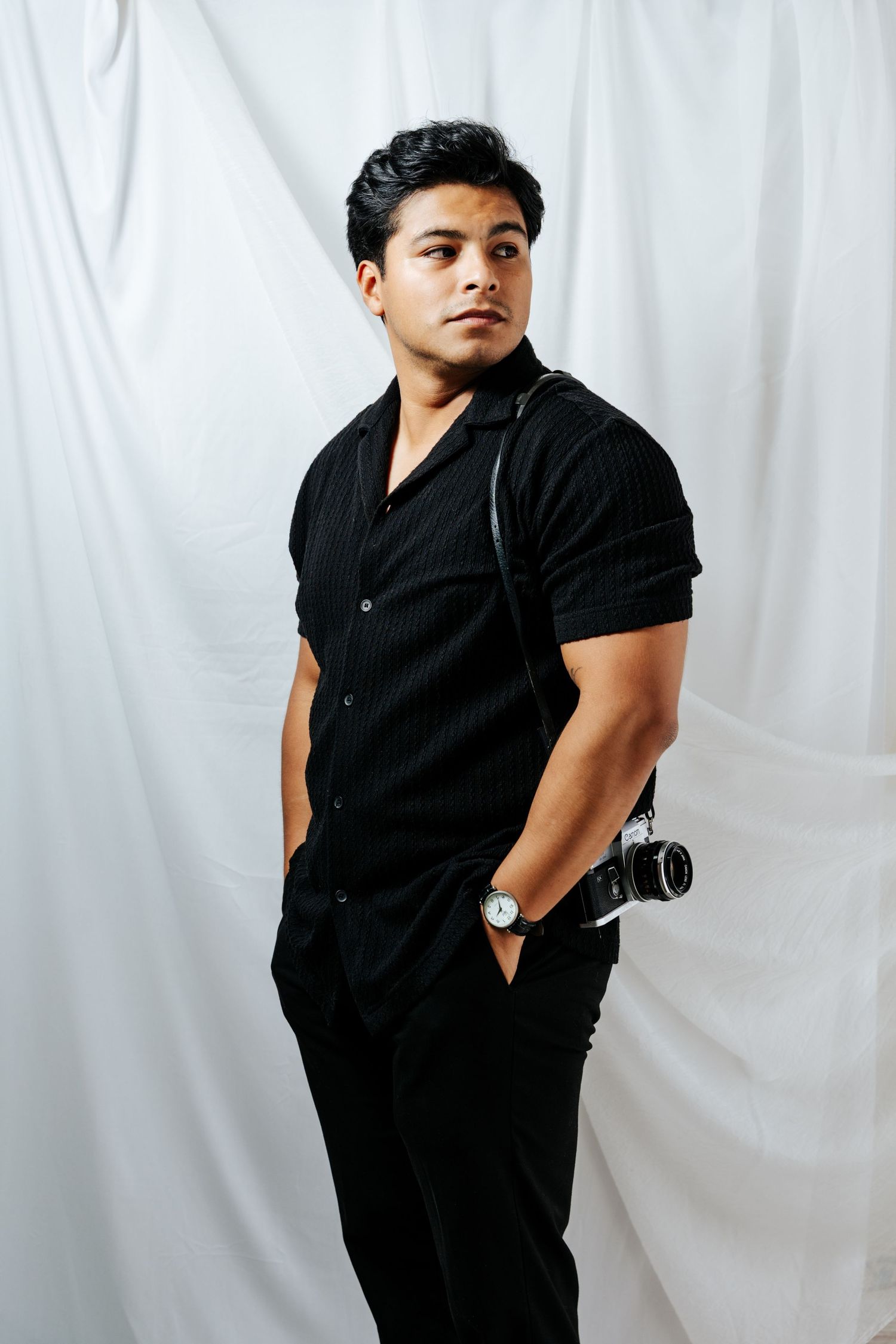 Young man in black shirt and pants holding a film camera on white backdrop studio portrait.