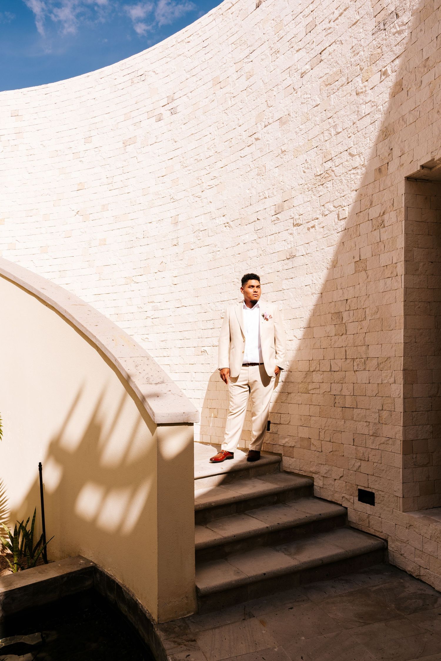 Groom in a cream suit and brown shoes stands on stone steps beside a curved white brick wall in bright sunlight.