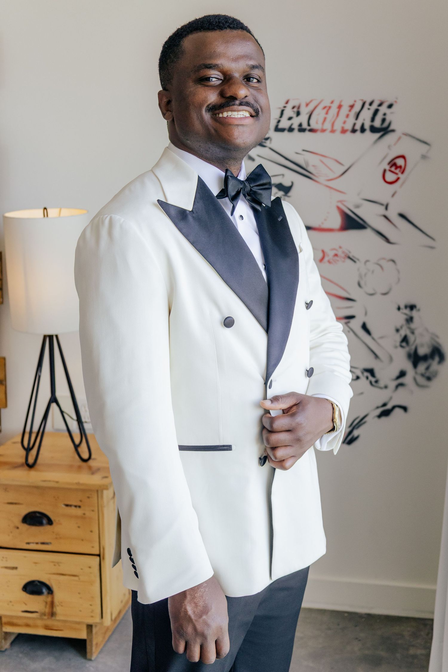 A person wearing a white tuxedo jacket with black lapels and bow tie poses in front of a wall with artwork.