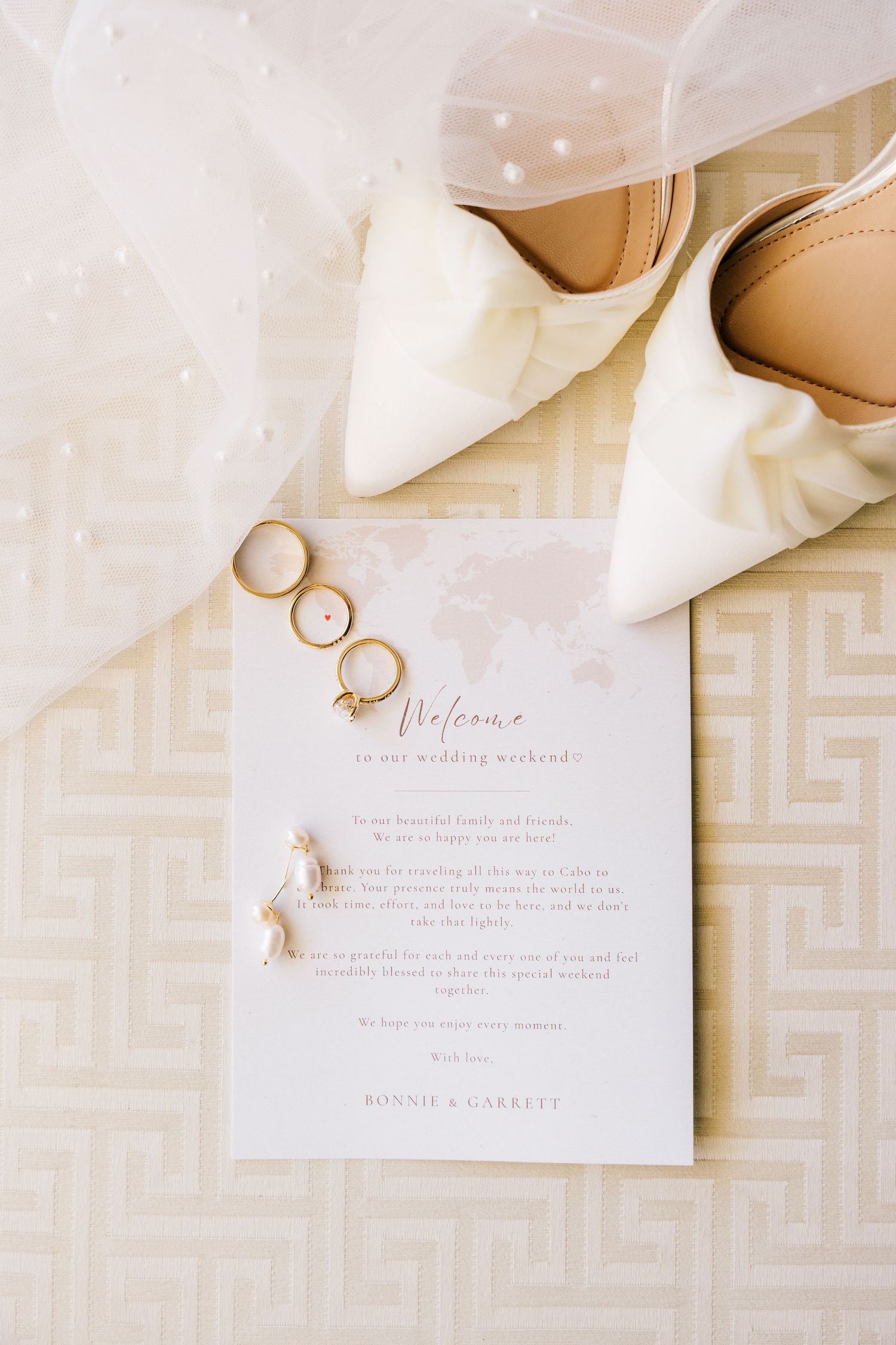 Wedding flat lay featuring gold rings, white heels, veil, and welcome card signed by Bonnie and Garrett on cream background.