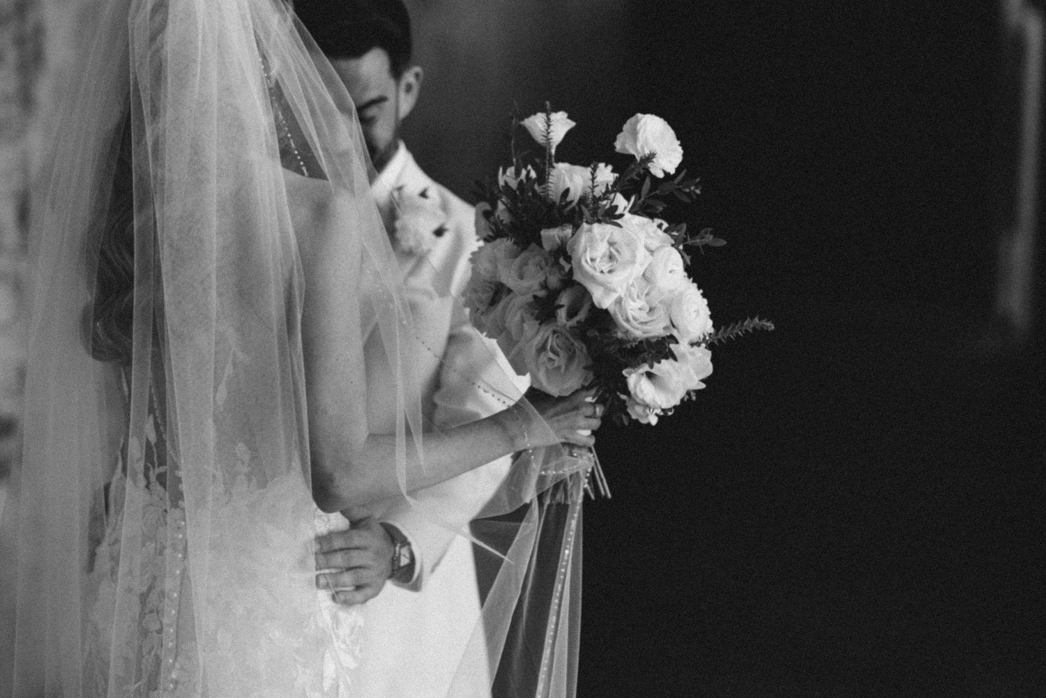 Black and white wedding photograph showing bride in elegant lace gown holding bouquet while standing by window light.