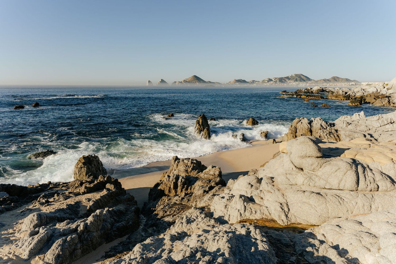Rocky coastline with crashing waves, sandy beach, and mountain views under clear blue sky in Cabo San Lucas, Mexico.